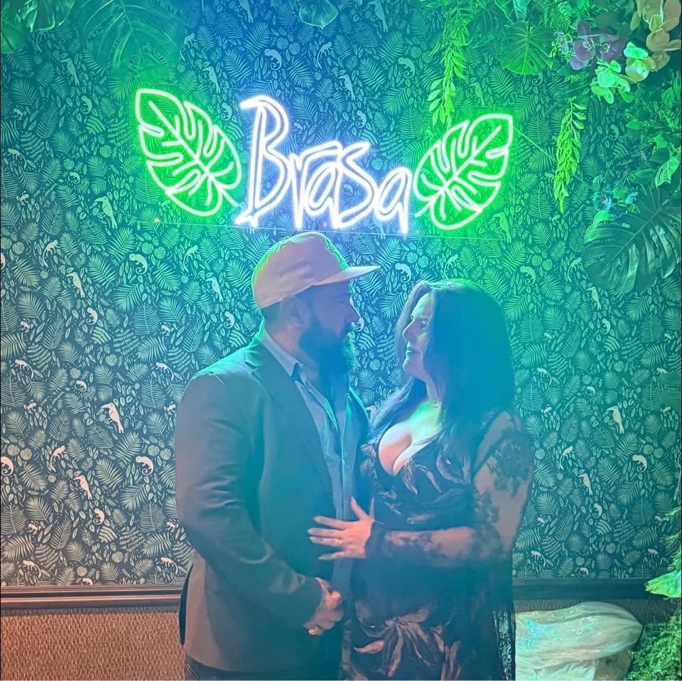 A man wearing a suit and a cap and a woman in a black lace dress are standing close together, gazing into each other's eyes. Behind them, there is a neon sign that says 'Brasil' surrounded by green leaves, and the background is decorated with a leafy tropical pattern.