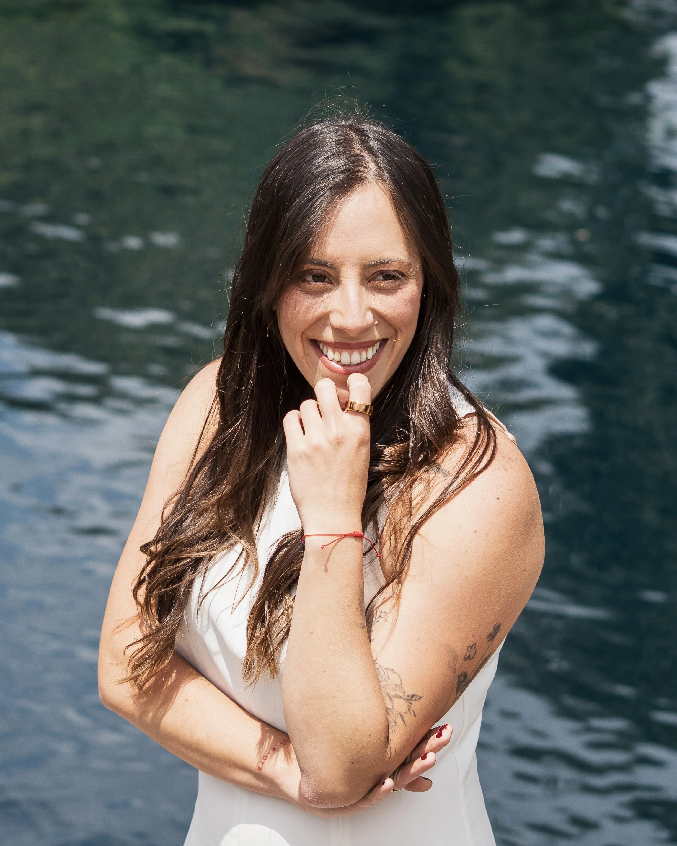 Mujer con cabello castaño largo, sonriendo, con fondo de agua, usando una blusa blanca, con tatuajes en el brazo, y accesorios en las manos y muñecas.