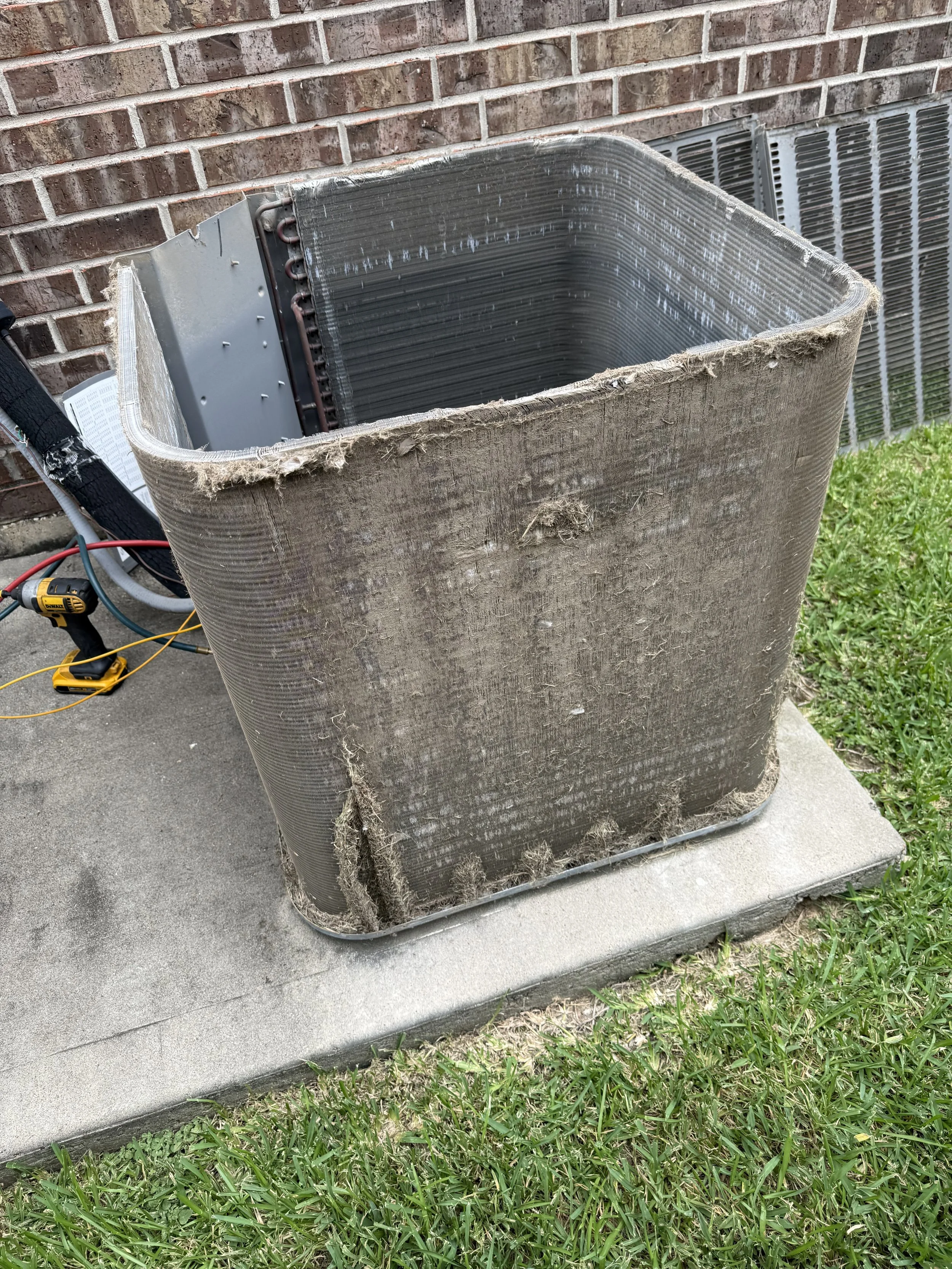 An HVAC unit's outer cover is removed, revealing the internal coils and components. The unit sits on a concrete pad adjacent to a brick wall and some grass.