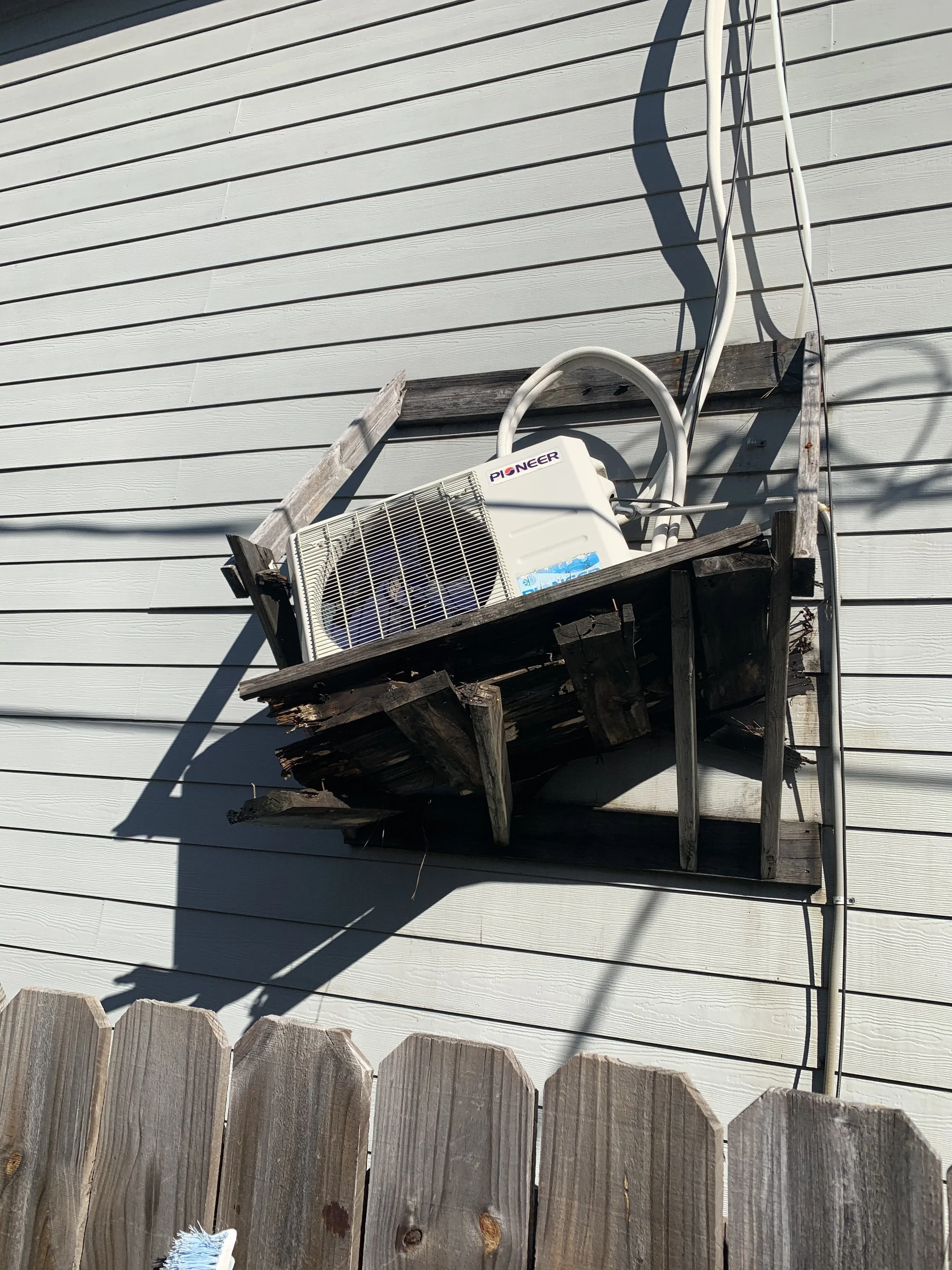 Wooden balcony with an air conditioning unit, electrical cables, and a weathered wooden fence in front of a house with white horizontal siding.
