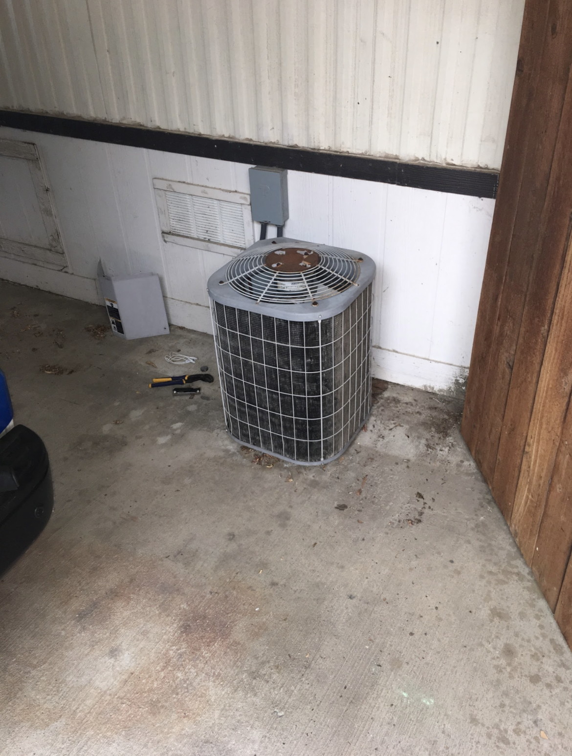 An outdoor area with a large, rusted air conditioning unit against a white wall. Tools and miscellaneous items scattered on the concrete floor.