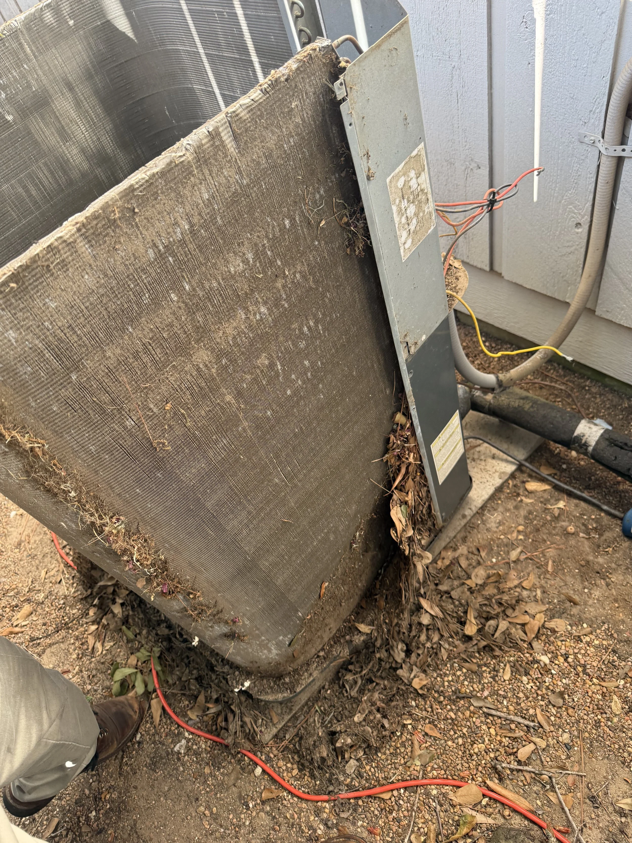 Dirty HVAC or air conditioning unit with visible wires and dirt buildup, placed on soil and surrounded by dry leaves.
