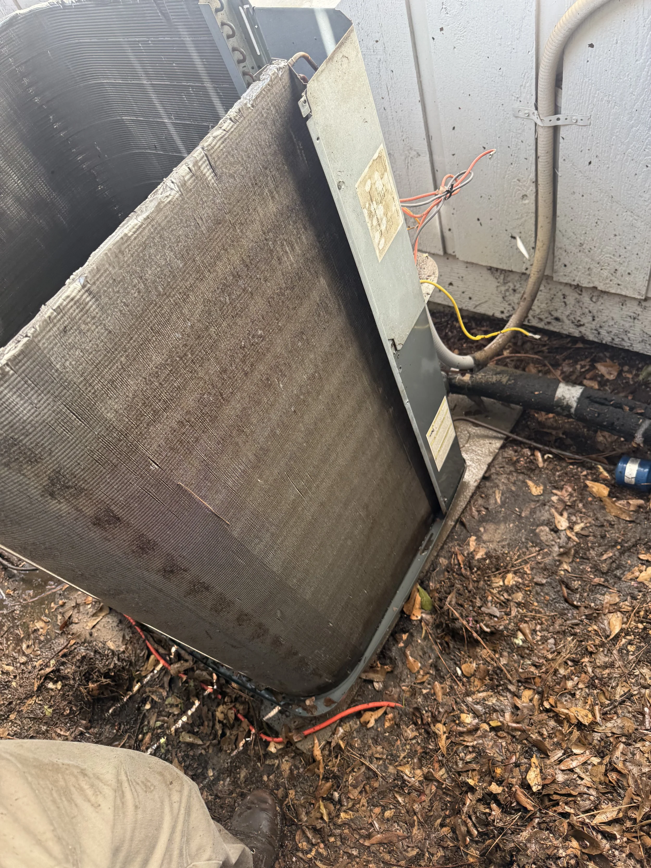 An outdoor scene showing a disassembled air conditioning or heat pump unit on the ground with exposed wiring and a dirty outdoor wall in the background, surrounded by fallen leaves.