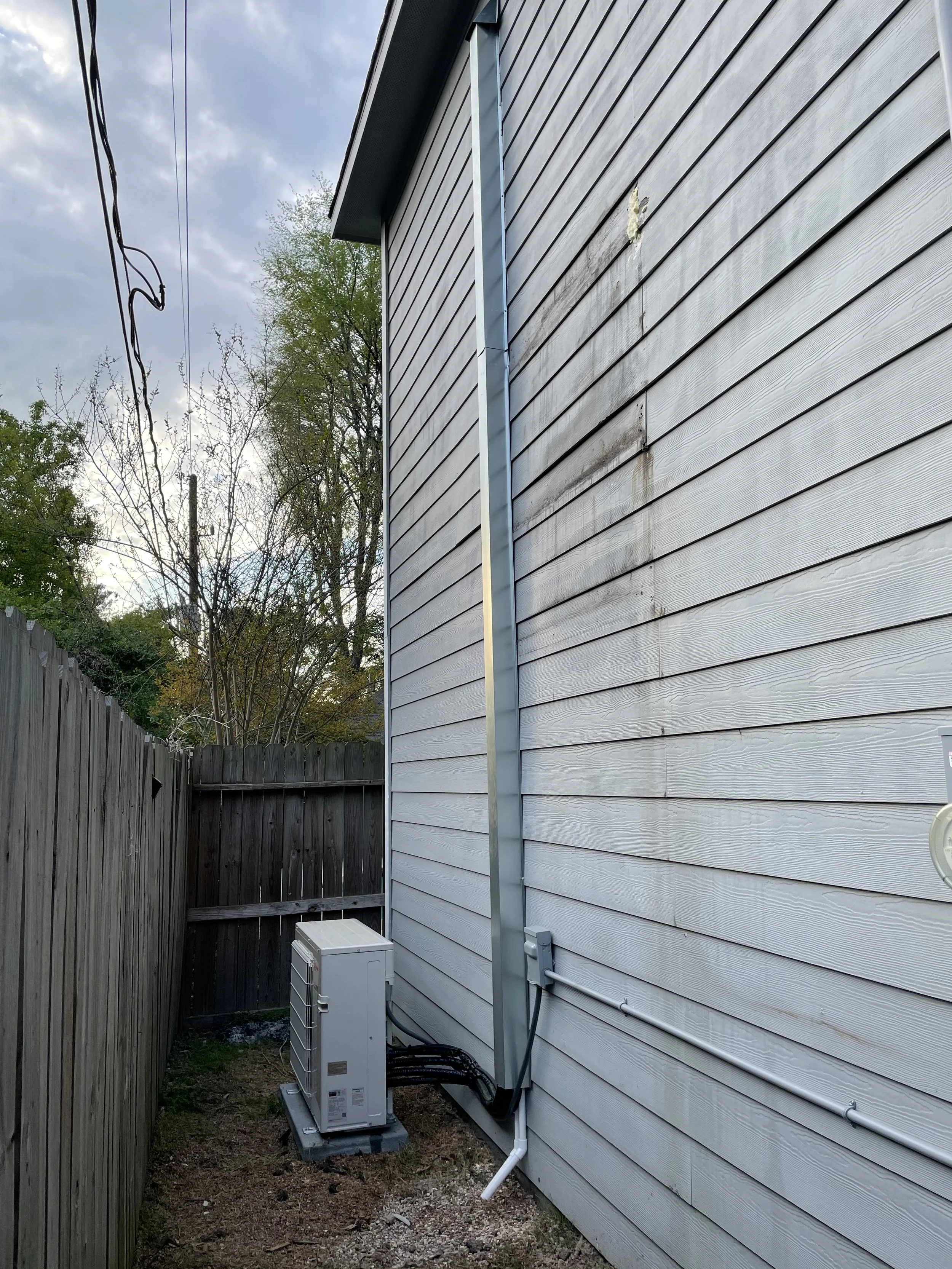 Side of a house with HVAC unit, electrical box, and visible wiring. Wooden fence and trees in background.