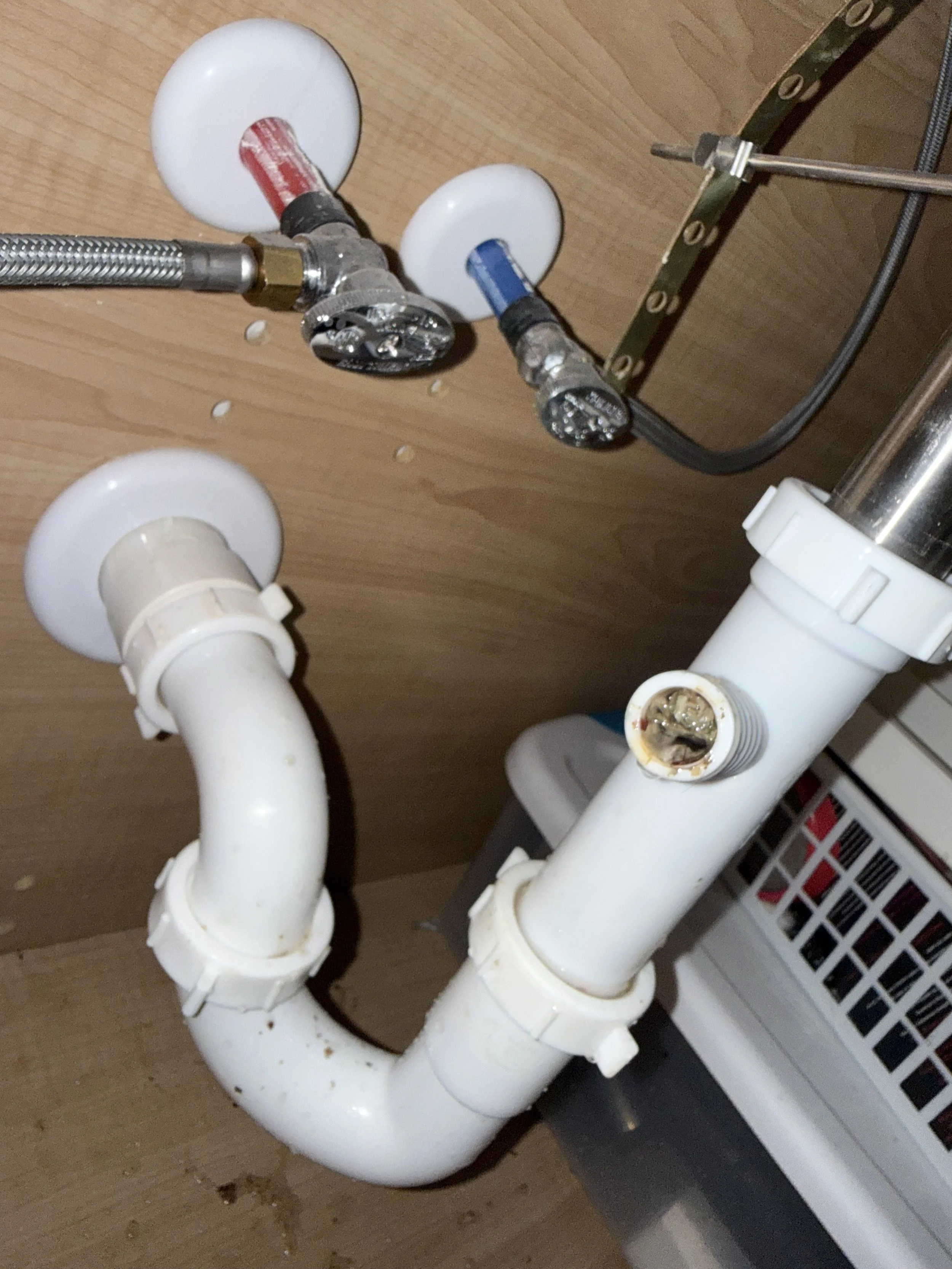 Underneath view of plumbing pipes and fittings, including PVC pipes and valve connections, attached to a wooden surface.