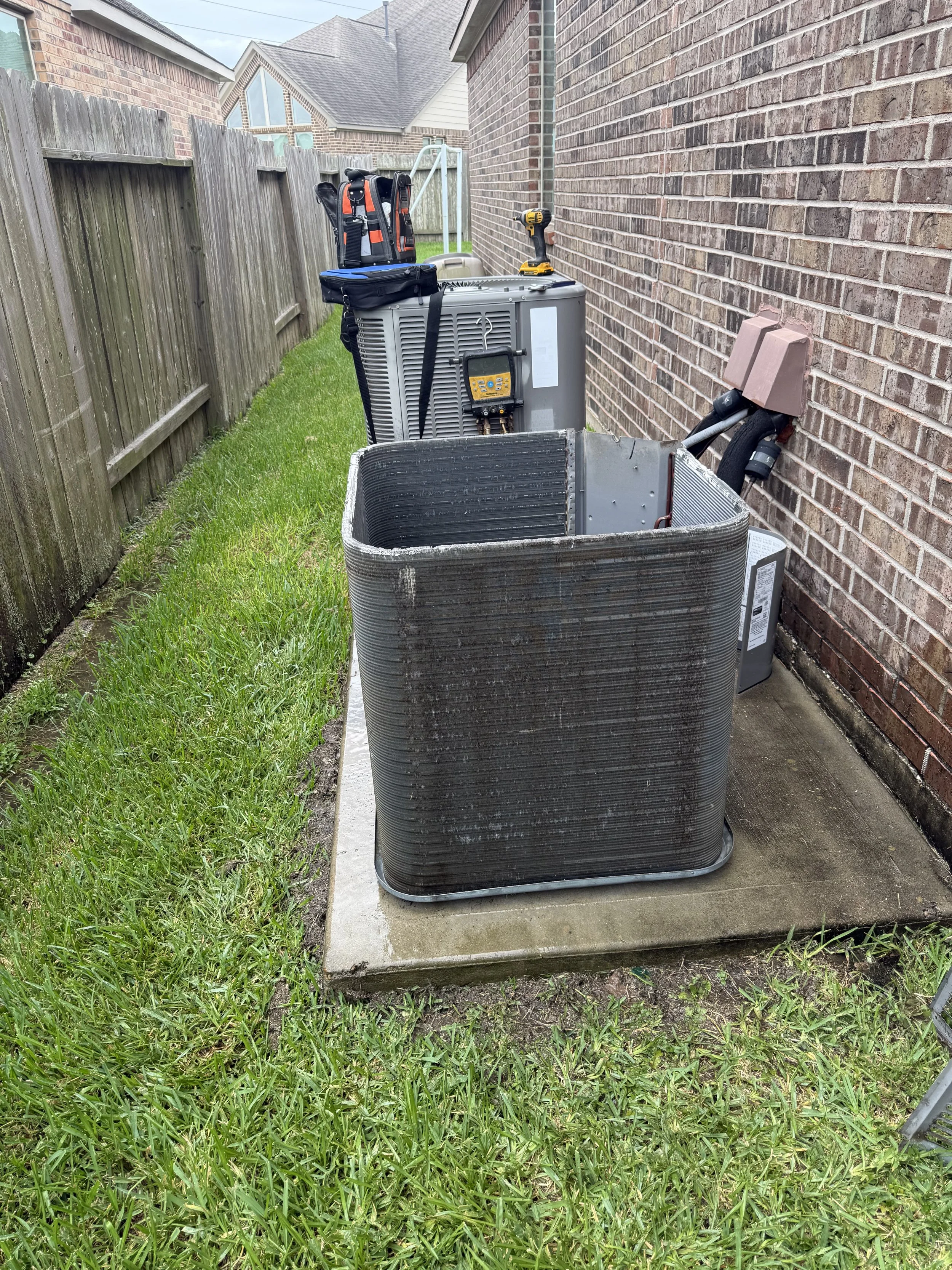 Outdoor air conditioning unit on concrete pad beside brick house wall, with tools and equipment on top, surrounded by grass and a wooden fence.