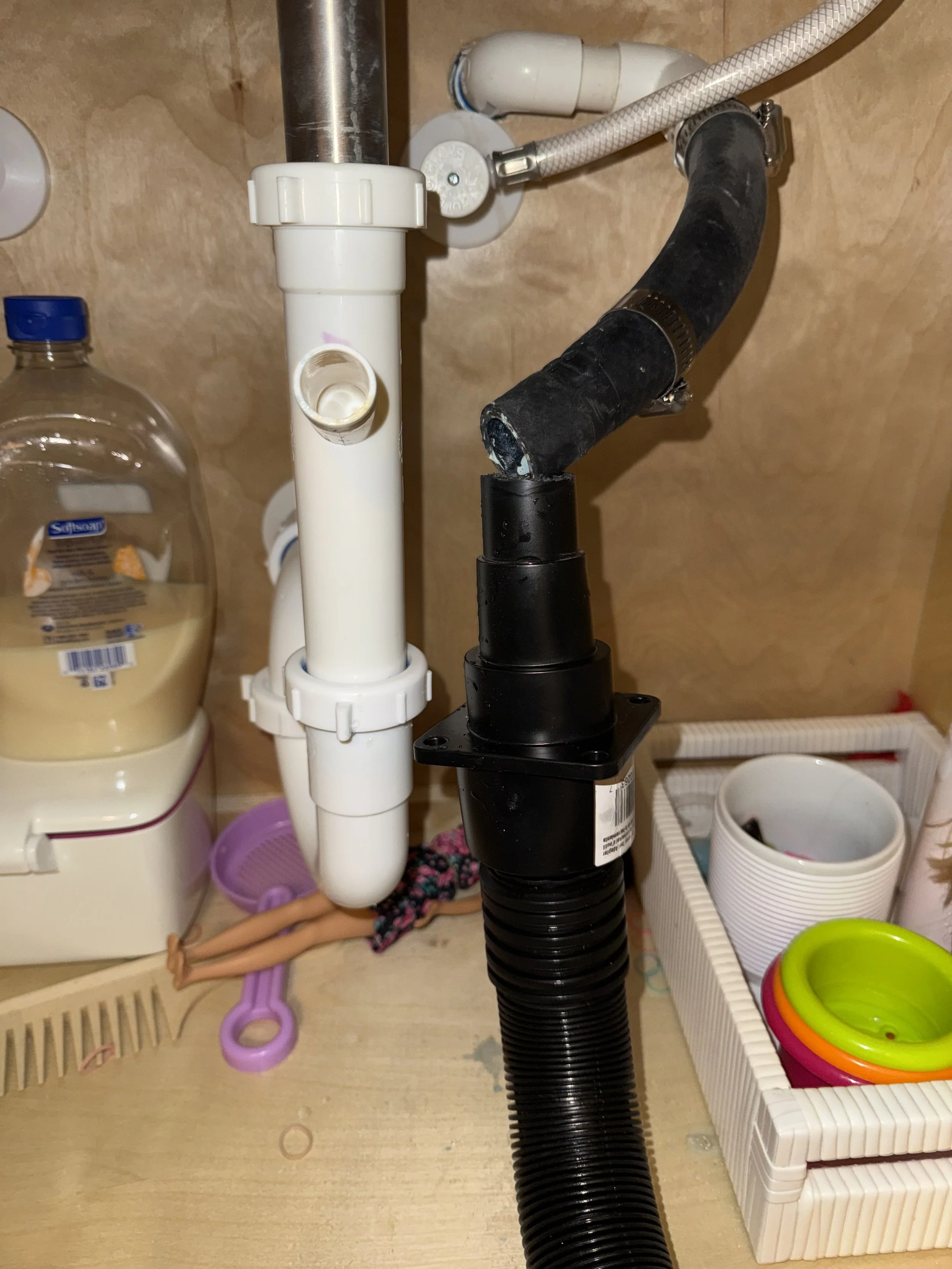 Under kitchen sink pipes with a black flexible hose and white PVC pipes, surrounded by cleaning supplies and kitchen utensils.