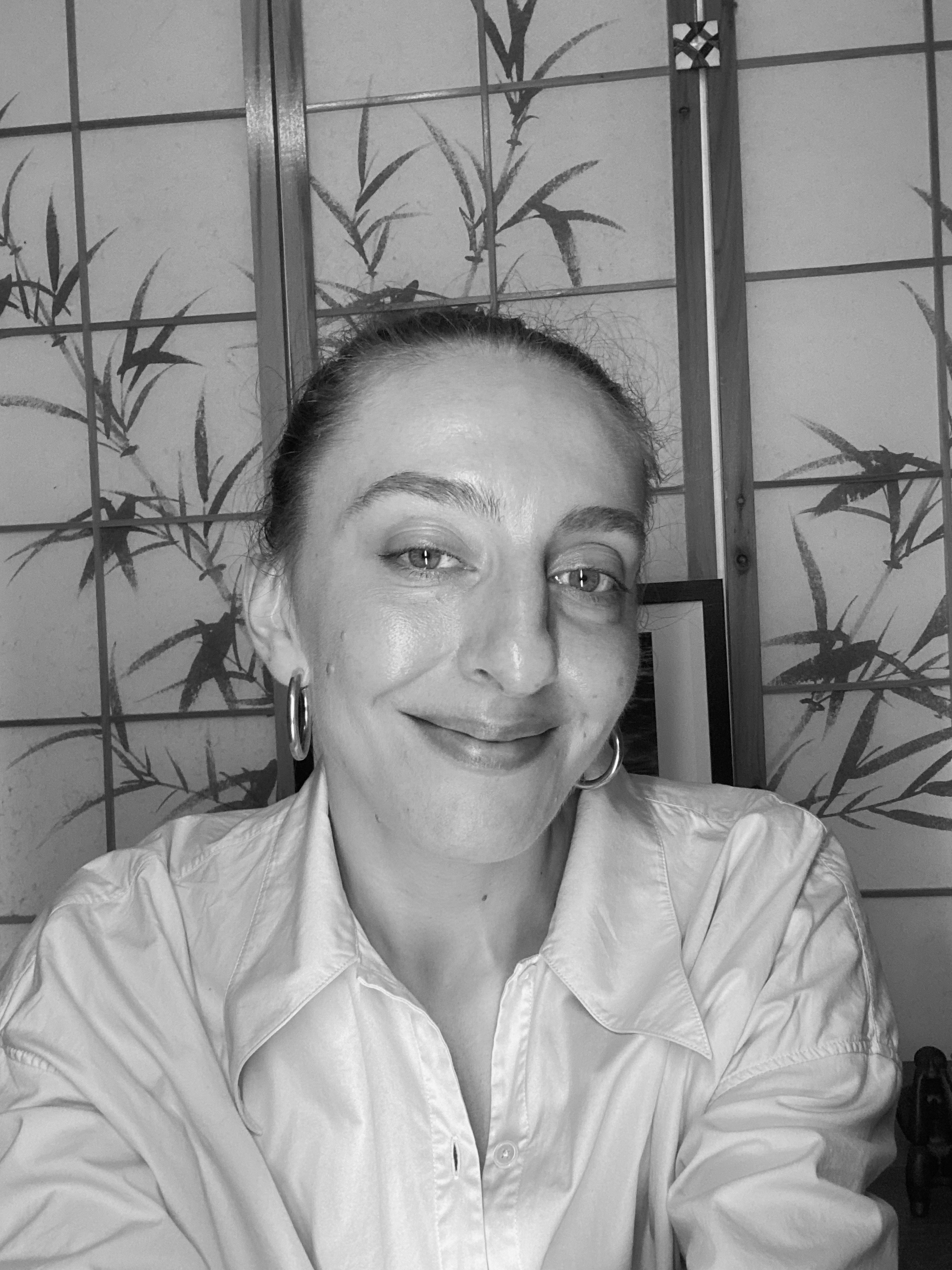 A woman with hair pulled back, smiling softly, wearing a light-coloured button-up shirt, sitting in front of a decorative bamboo-themed room divider in black and white.