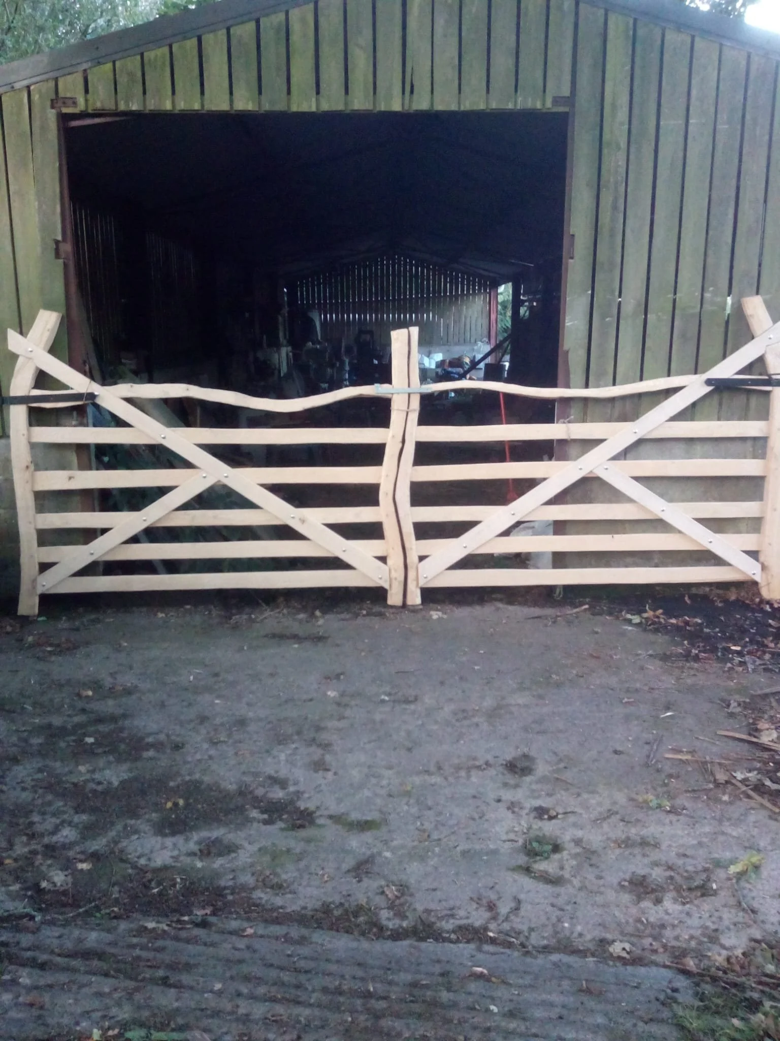 A wooden gate in front of a barn or shed. The gate is unfinished and made of horizontal wooden slats and diagonal supports. The barn has vertical wooden siding and is open, revealing some equipment and tools inside. The ground in front is dirt with some leaves and small plants.