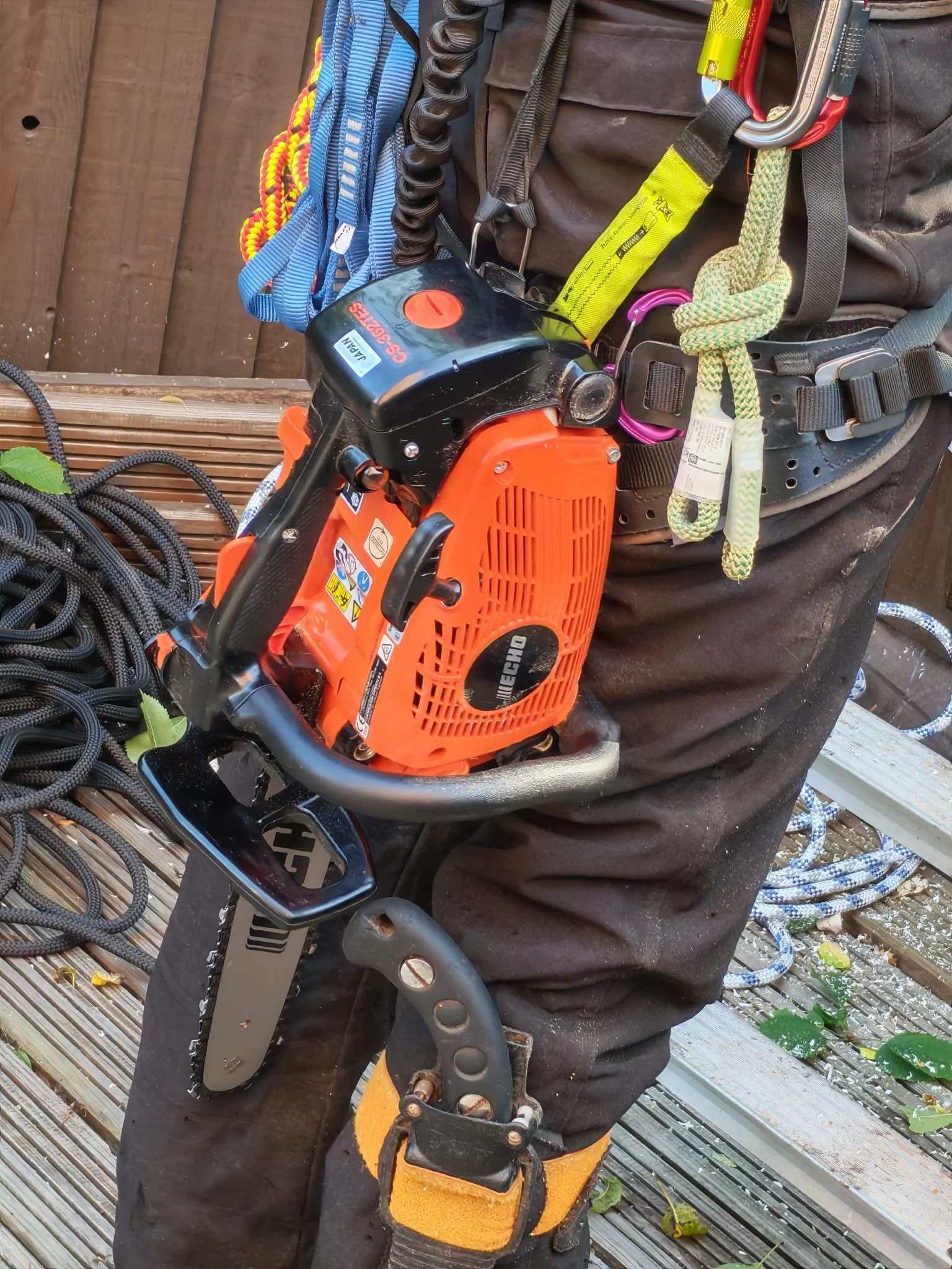 A person wearing black pants with various climbing gear attached, including a small orange chainsaw, climbing harnesses, carabiners, ropes, and a climbing helmet, standing on a wooden deck.