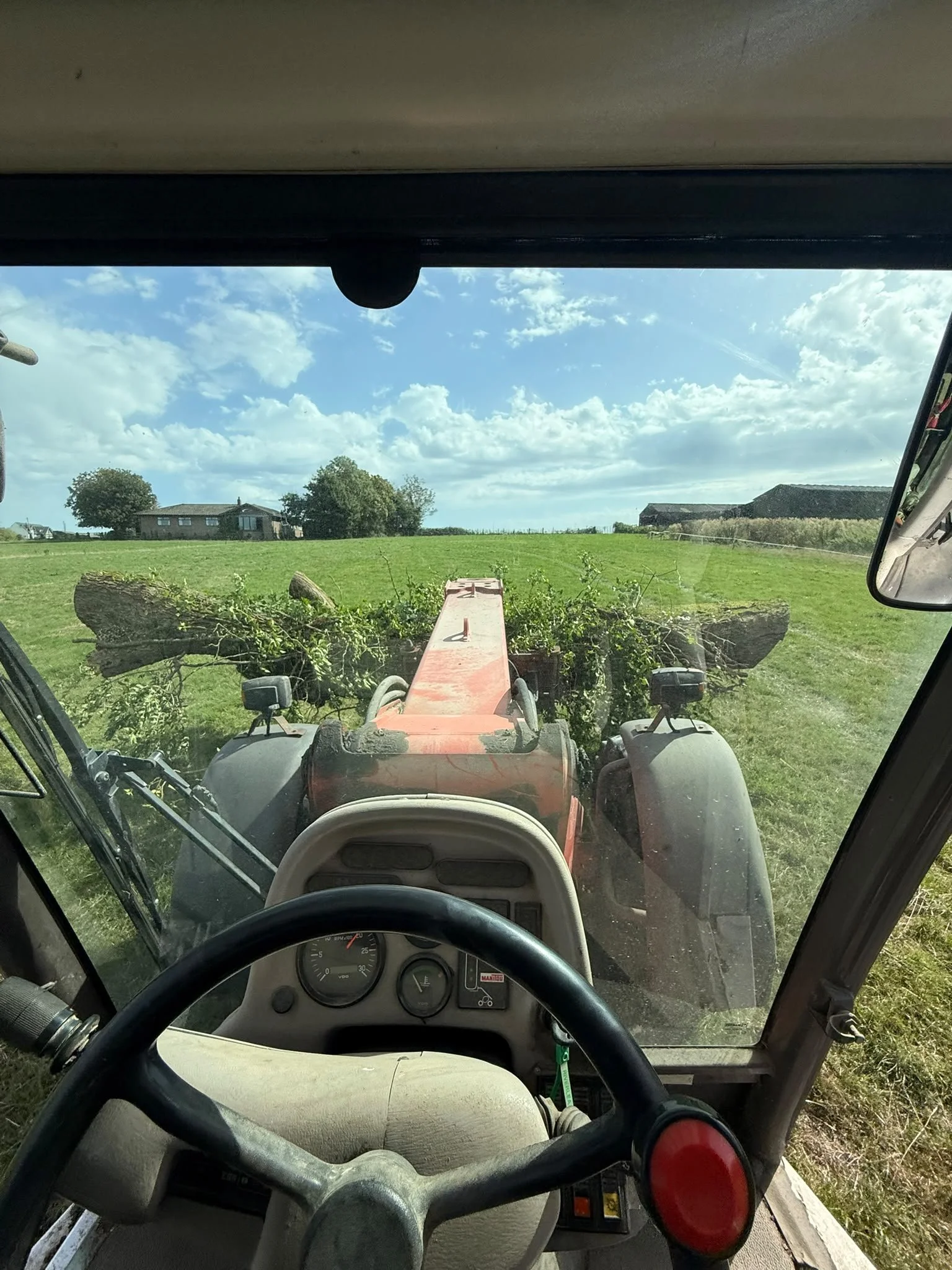View from inside a tractor cab looking through the front windshield at a green field, trees, farm buildings, and a blue sky with white clouds.
