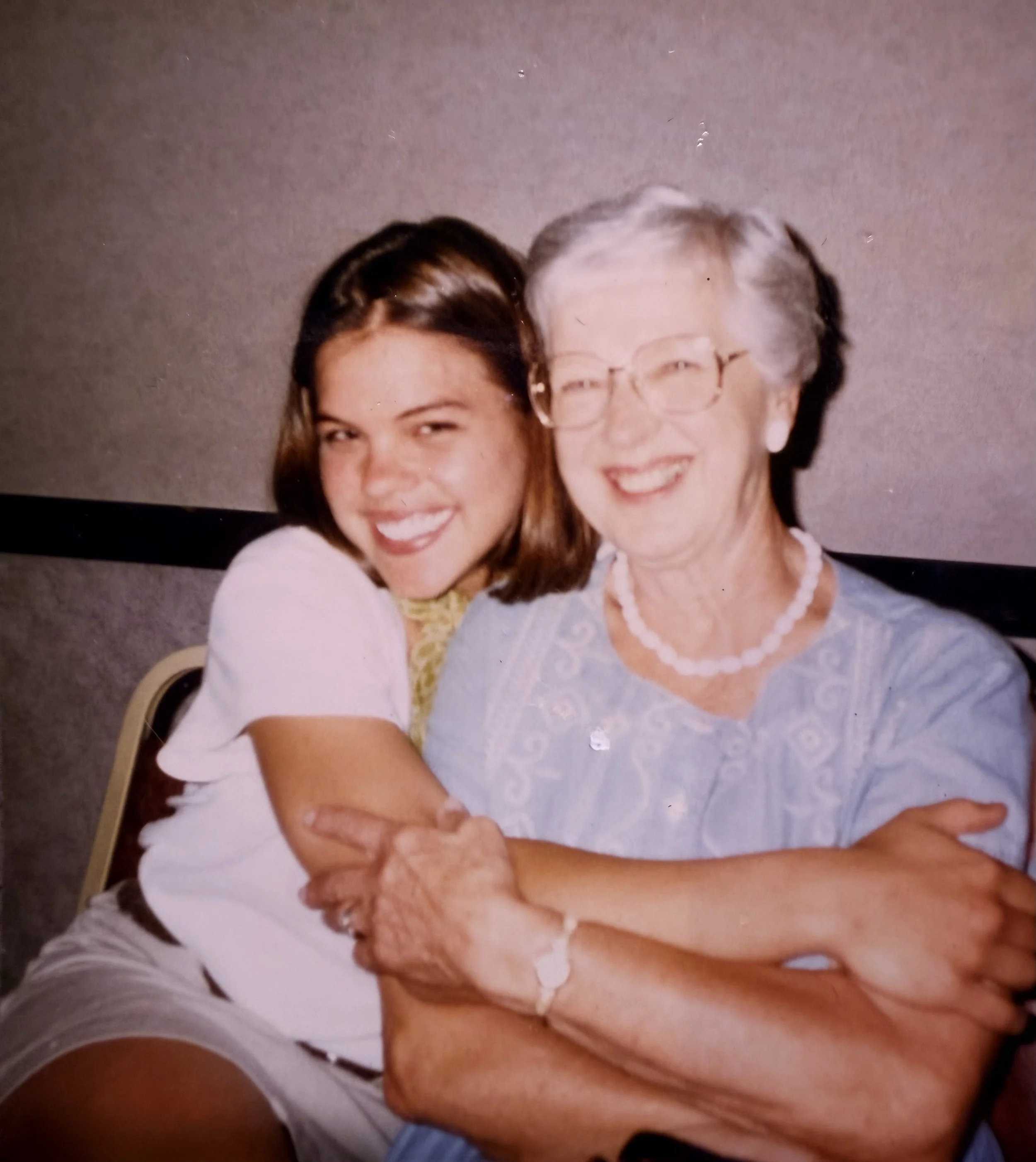 A woman and an elderly woman smiling together in a close-up photo, with the younger woman on the left and the older woman on the right, wearing glasses and a necklace, both dressed in light-colored tops. This is Mabel.