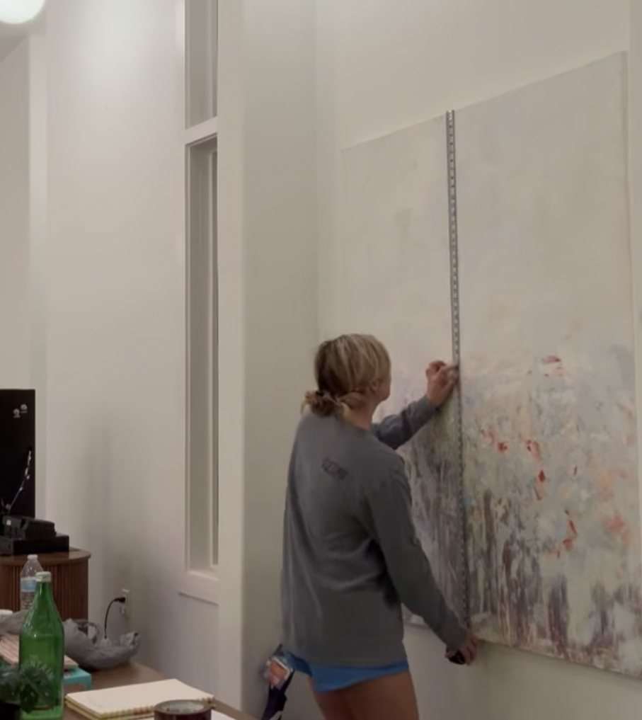 Person measuring a large abstract painting on a wall using a measuring tape in a room with minimal decor.