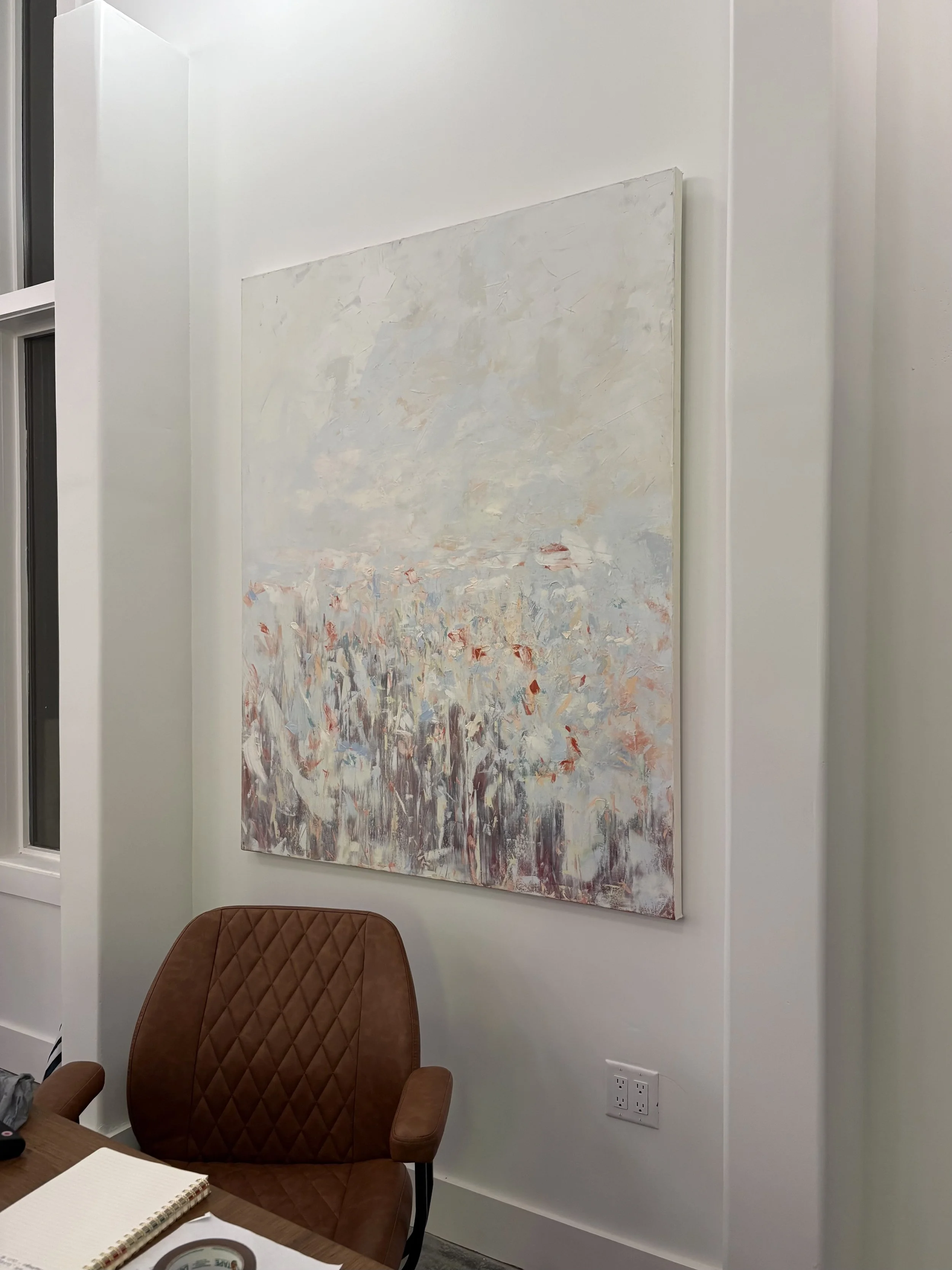 A modern office with a white wall, a large abstract painting, a brown quilted armchair, a desk with a notebook and a roll of tape, and electrical outlets on the wall.
