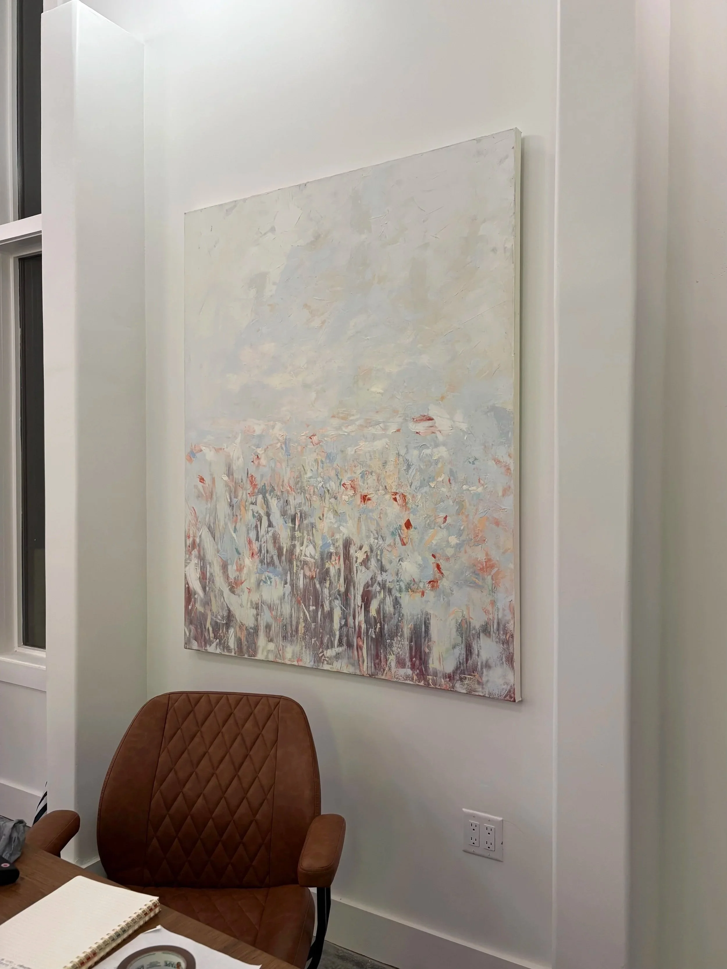 An office with a large abstract painting on a white wall, brown upholstered chair, and a desk with notebooks and a roll of tape.