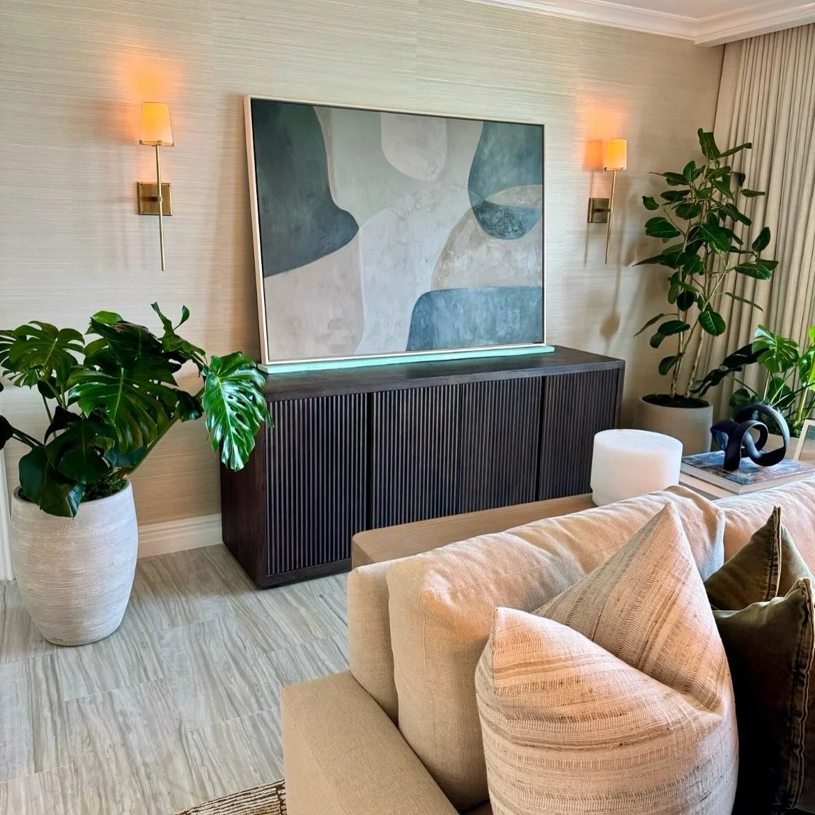 Living room with a beige sofa, large potted plants, abstract artwork on a dark wooden cabinet, and two wall-mounted lamps with beige shades.