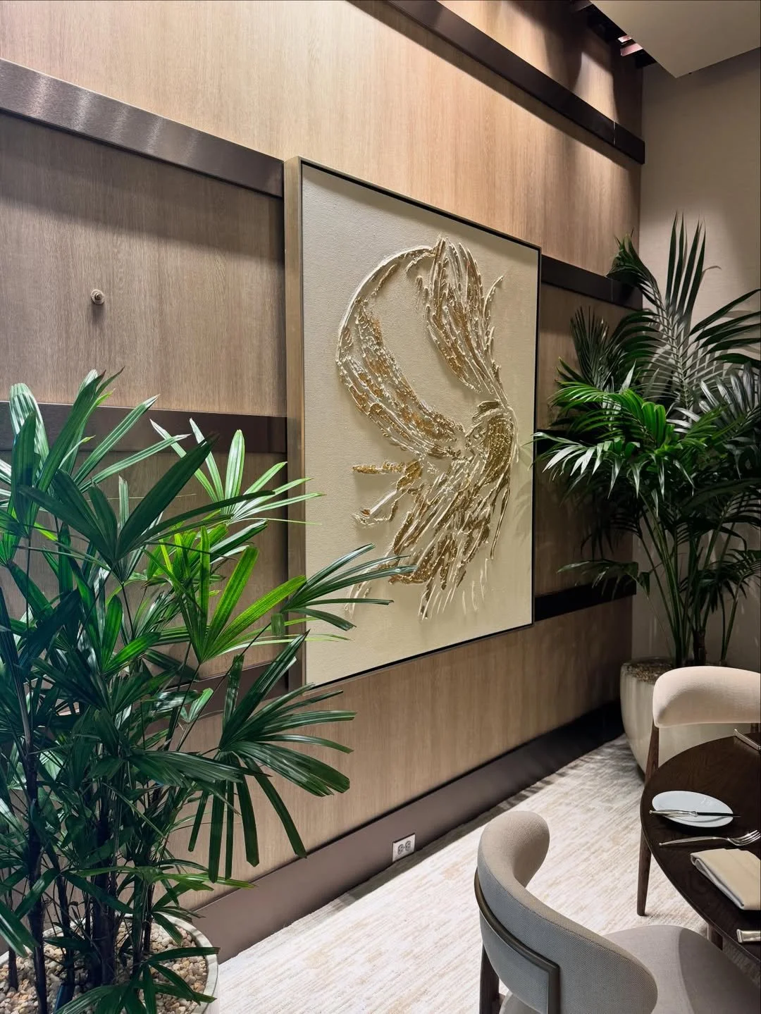Interior of a modern dining area with large green houseplants, a beige carpet, and a wall art piece depicting a bird in gold textures on a light-colored background.