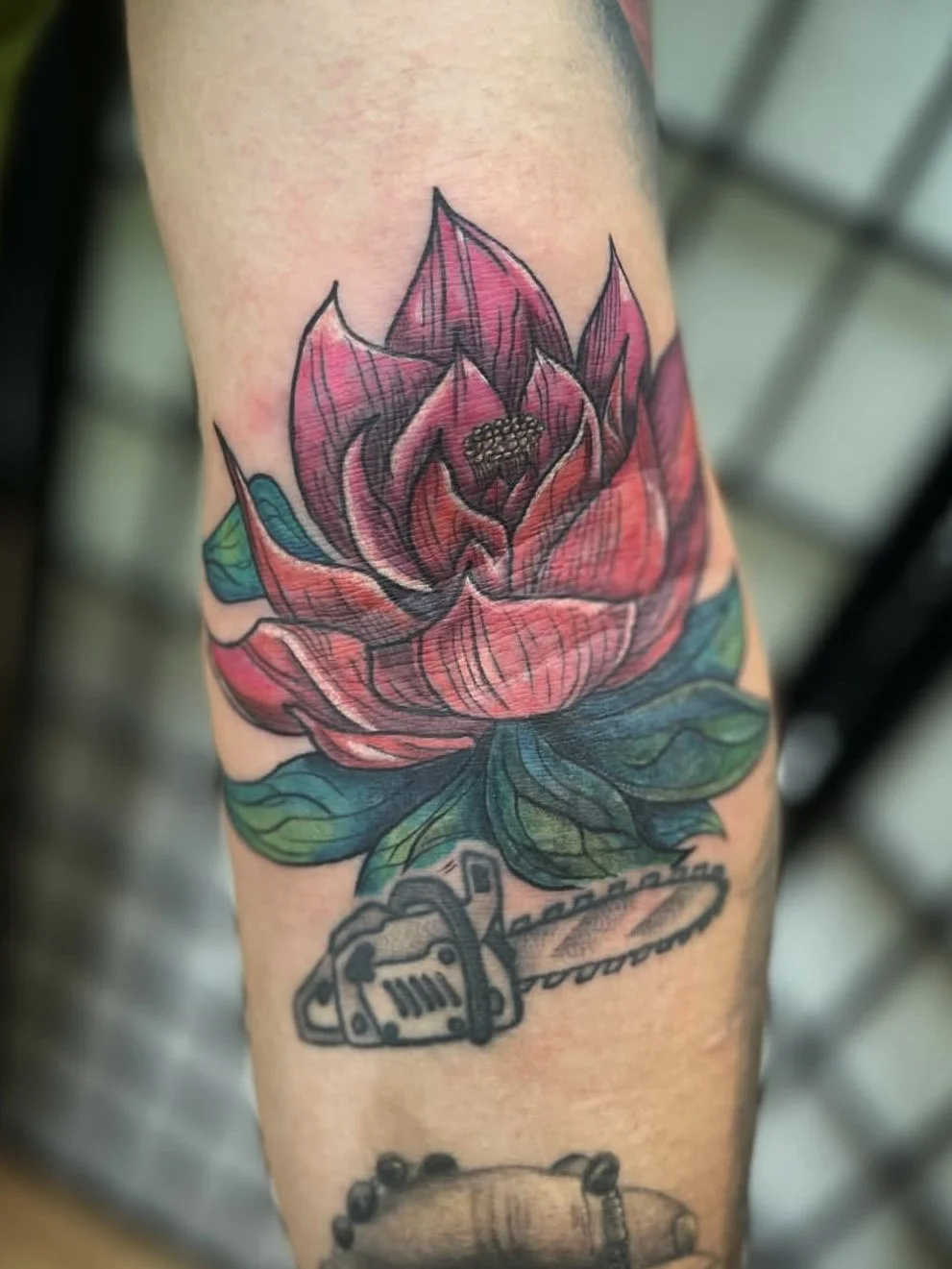 Colorful tattoo of a pink lotus flower with green leaves on arm, along with a black and gray chainsaw design below.