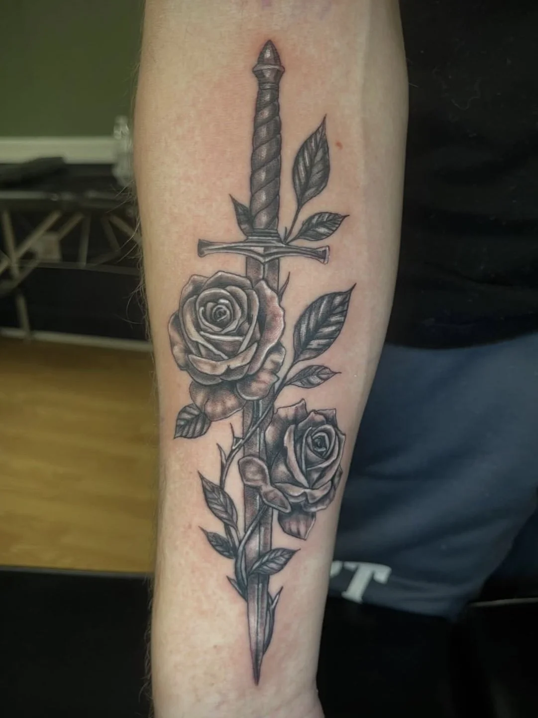 Tattoo of a sword with roses and leaves in black and gray ink on a person's forearm.