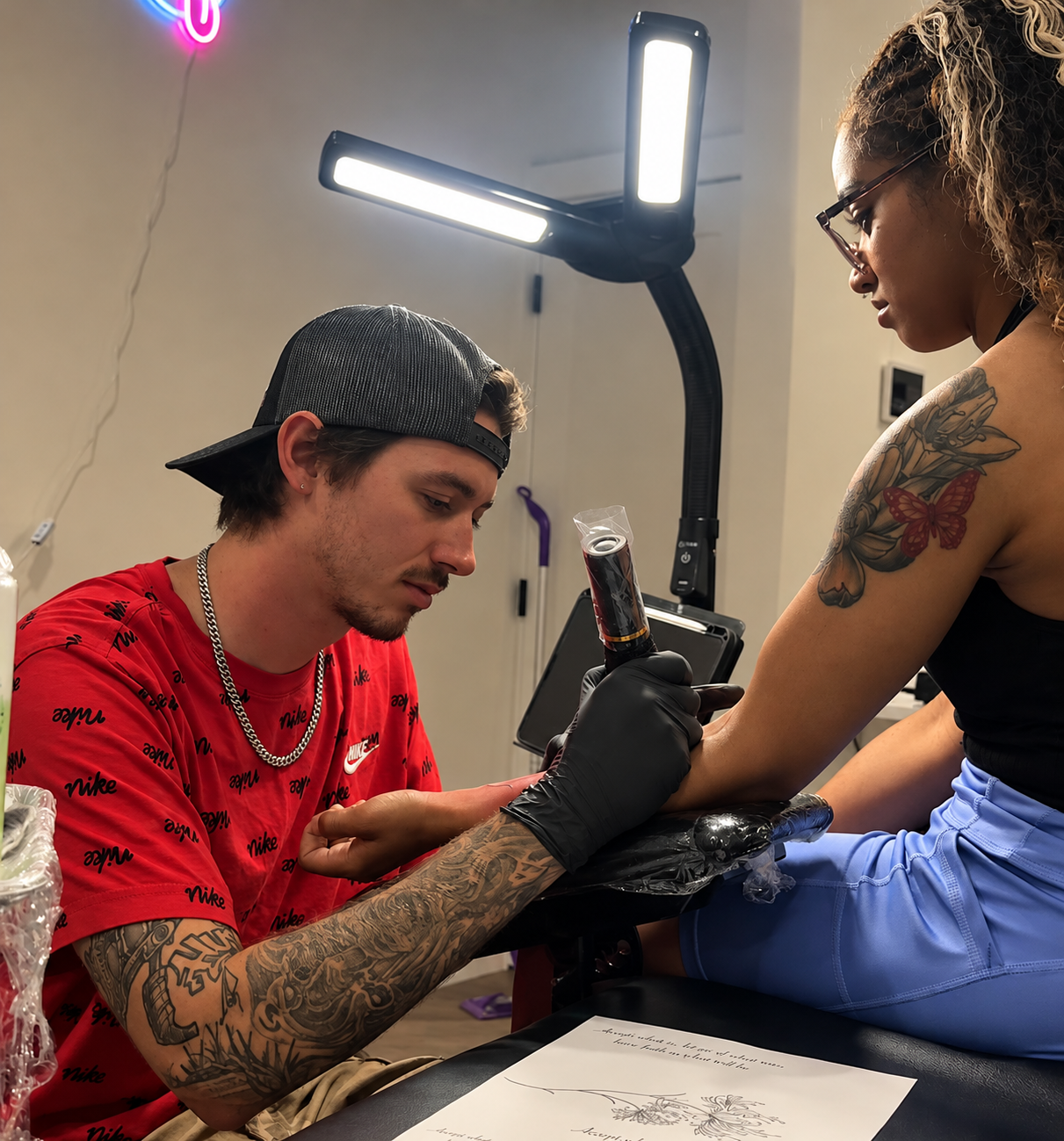 A tattoo artist with tattoos on his arms and wearing a red Nike T-shirt is tattooing a woman on her forearm. The woman has tattoos on her upper arm and is wearing glasses and a black sleeveless top. They are in a tattoo studio with professional lighting.