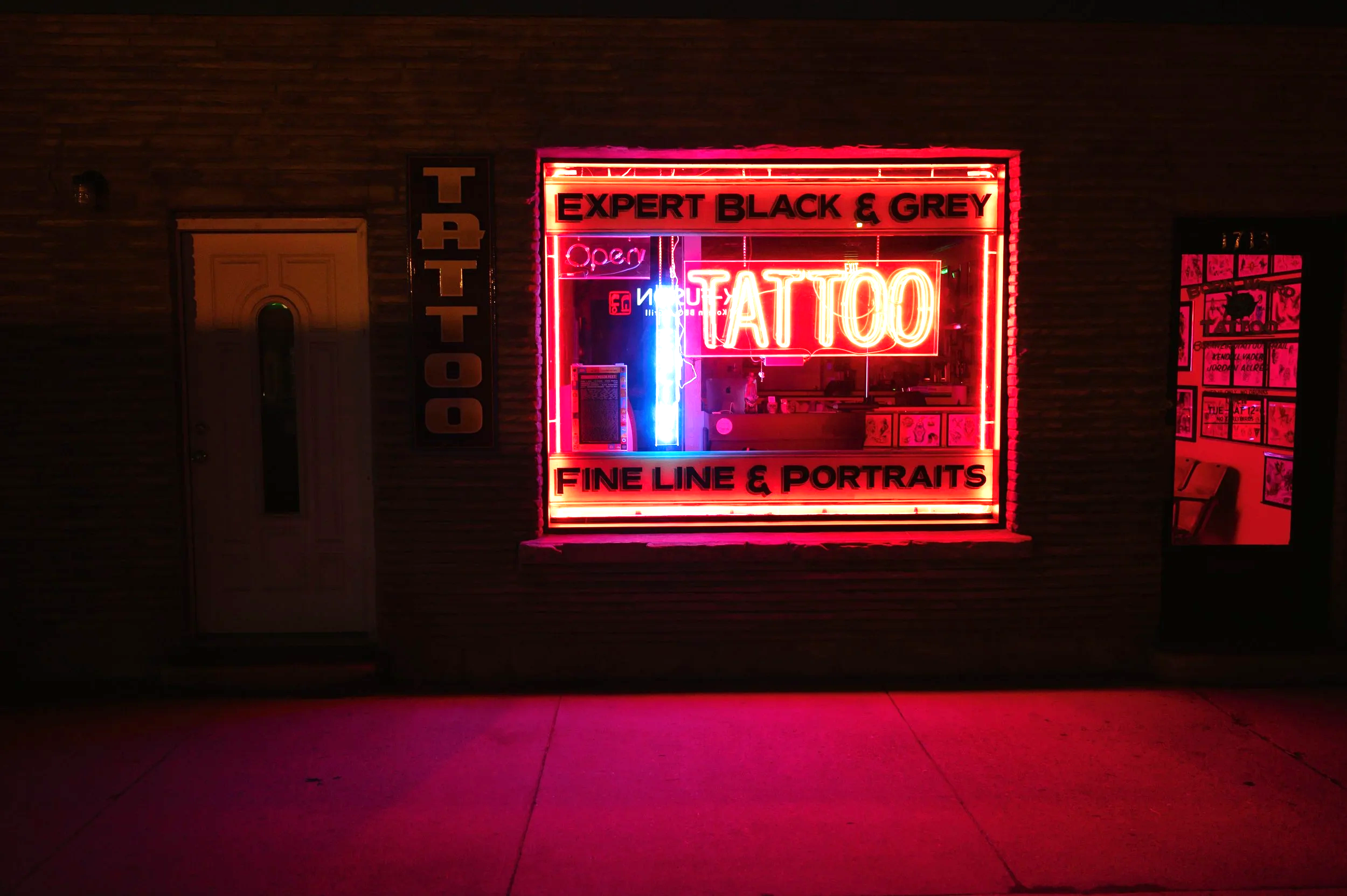 Bright neon sign outside a tattoo shop displaying the words "Expert Black & Grey," "Tattoo," "Piercing", and "Fine Line & Portraits," with a smaller "Open" sign and other neon symbols.