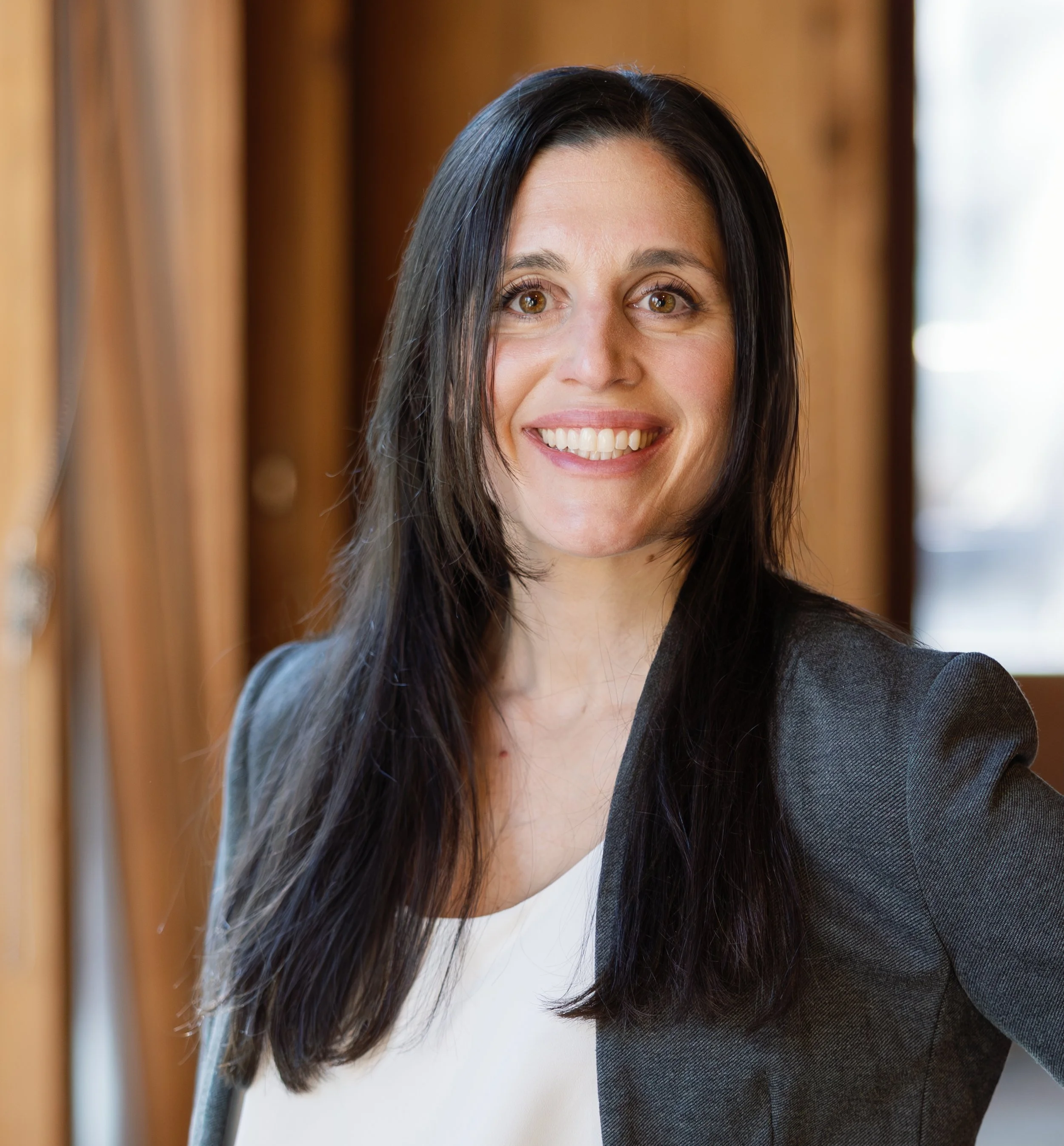 Close-up of a woman with long dark hair smiling, wearing a dark blazer and white shirt, indoors with warm wooden paneling and a natural light source.