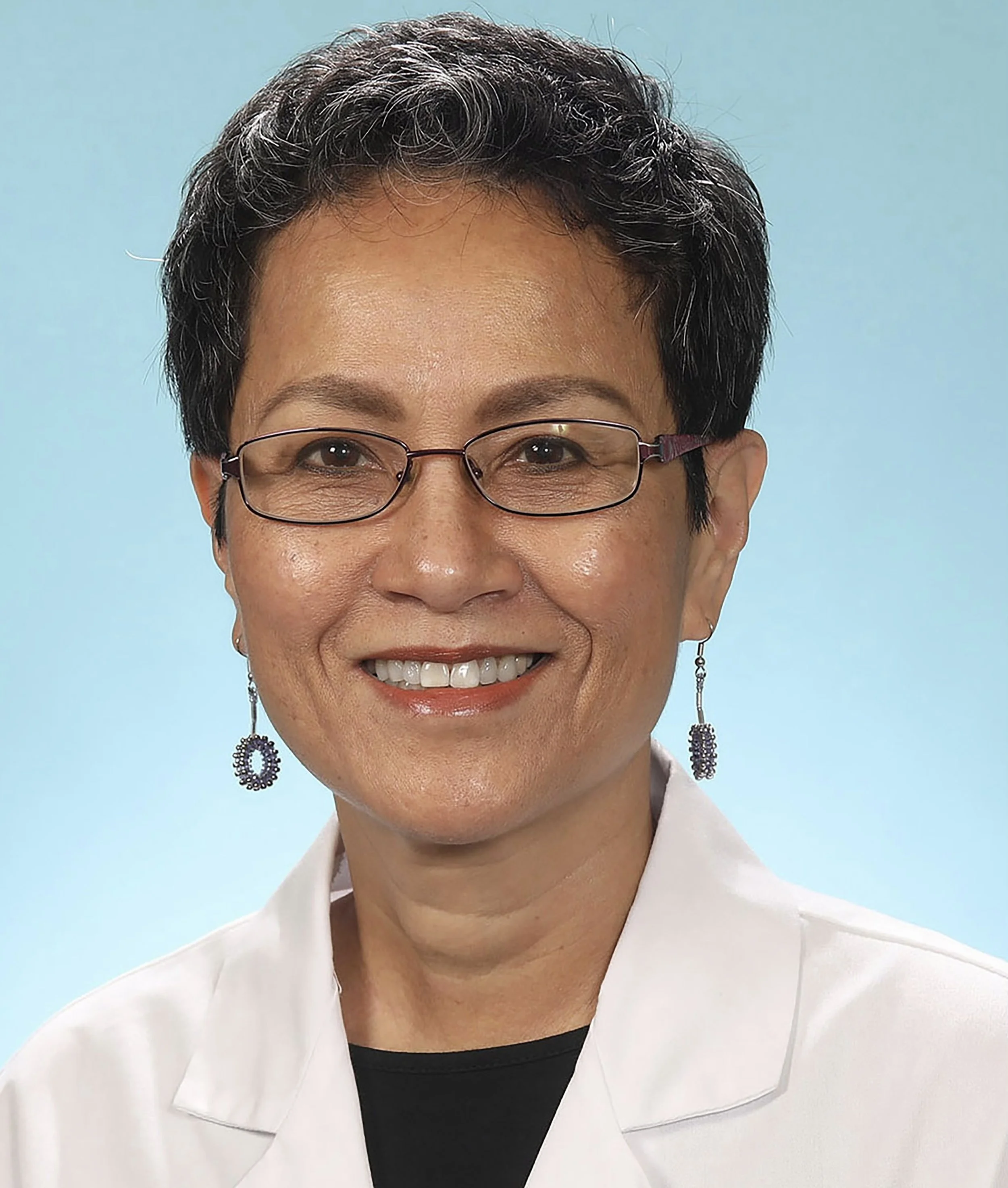 Close-up of a woman with short curly black hair wearing glasses, earrings, a white jacket, and a black top, smiling against a light blue background.