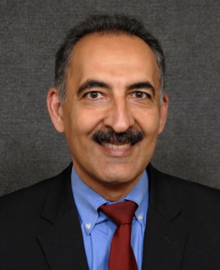 A smiling man with a mustache wearing a black suit, blue shirt, and red tie against a dark gray background.