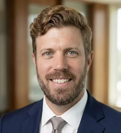A professional man with a beard and curly light brown hair, wearing a dark suit and light shirt, smiling in an indoor setting.