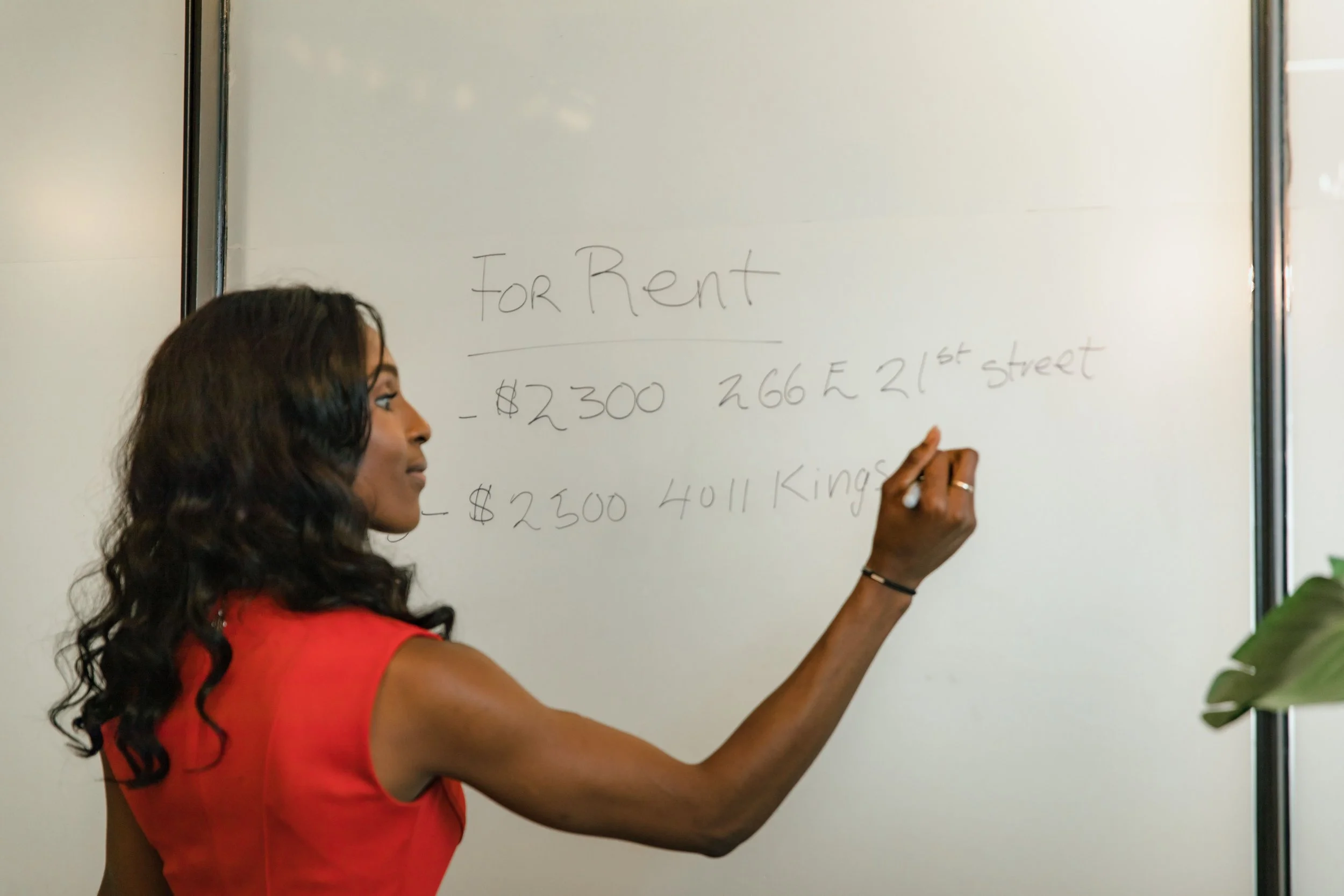 A woman with dark wavy hair wearing a red shirt writing on a whiteboard with words about rental prices.