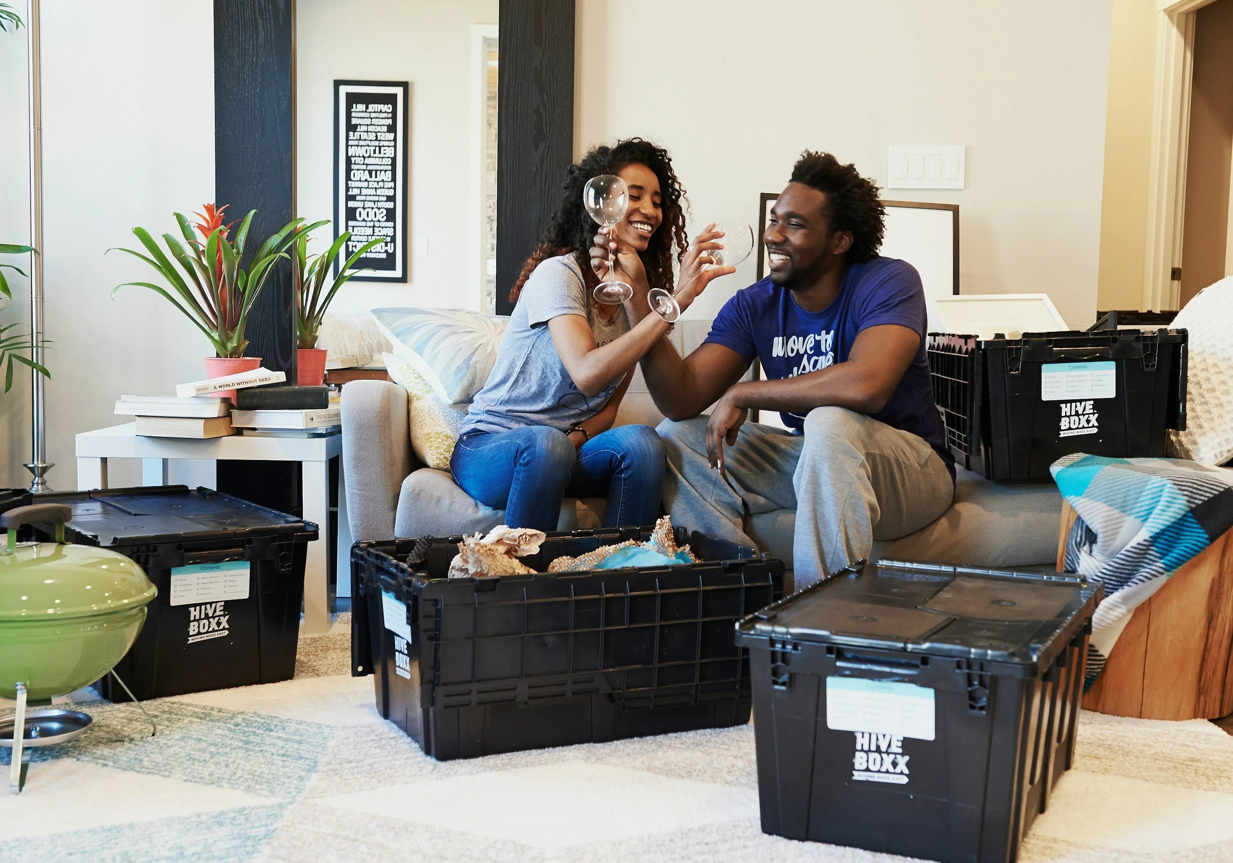 Two smiling people, a woman with curly hair and a man with an afro, sitting on a sofa, holding wine glasses, and giving each other a fist bump. There are multiple black plastic storage crates labeled 'HIVE BOXX' in the room, with some open and others stacked. The background includes a framed poster on the wall, potted plants on a white side table, and a few books.