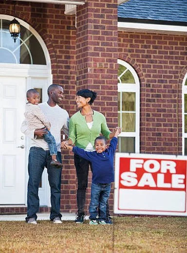 Family standing outside a house with a for sale sign, including two children, a mother, and a father.