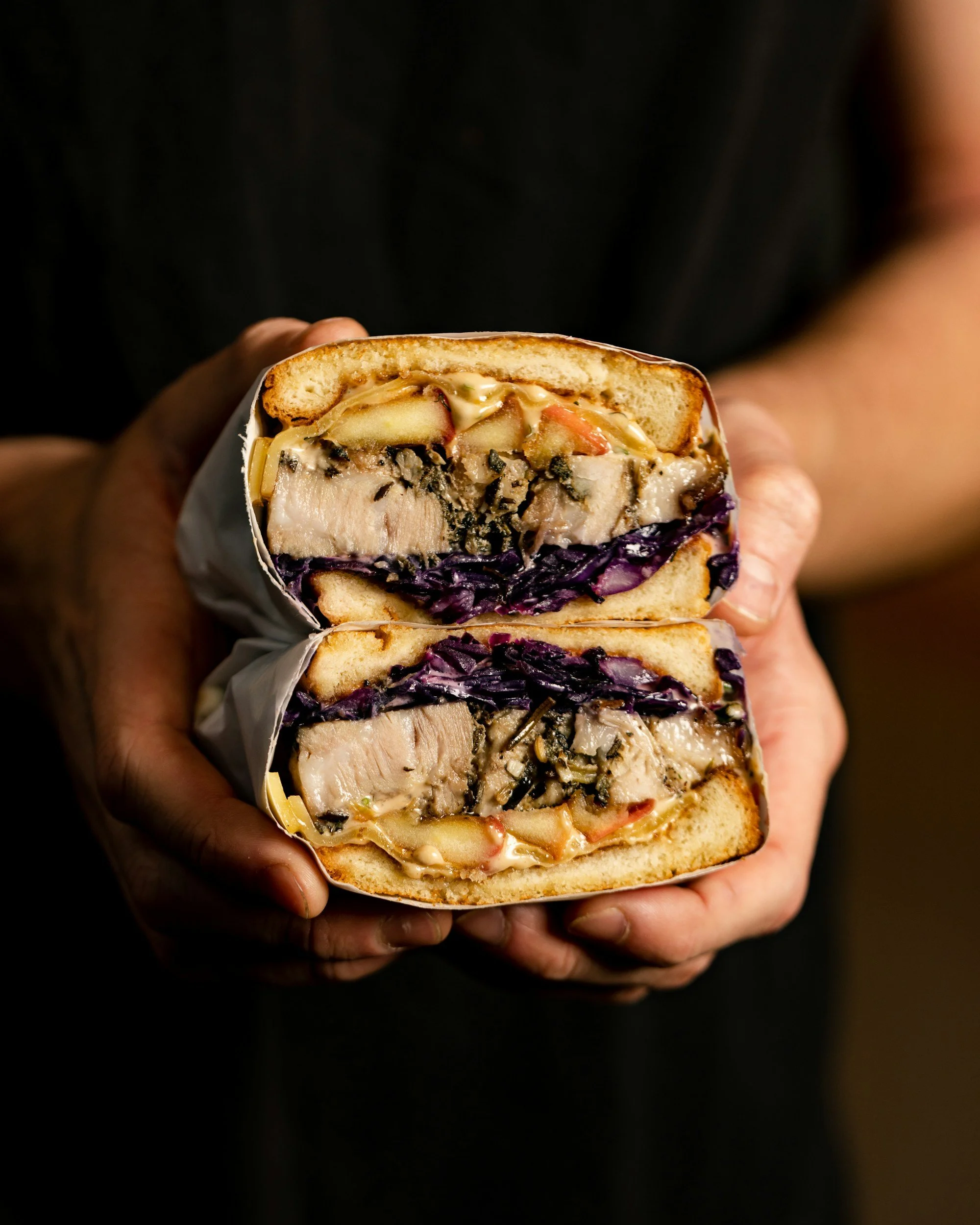 Close-up of a hand holding a sandwich with layers of bread, meat, vegetables, and sauces, with a dark background.