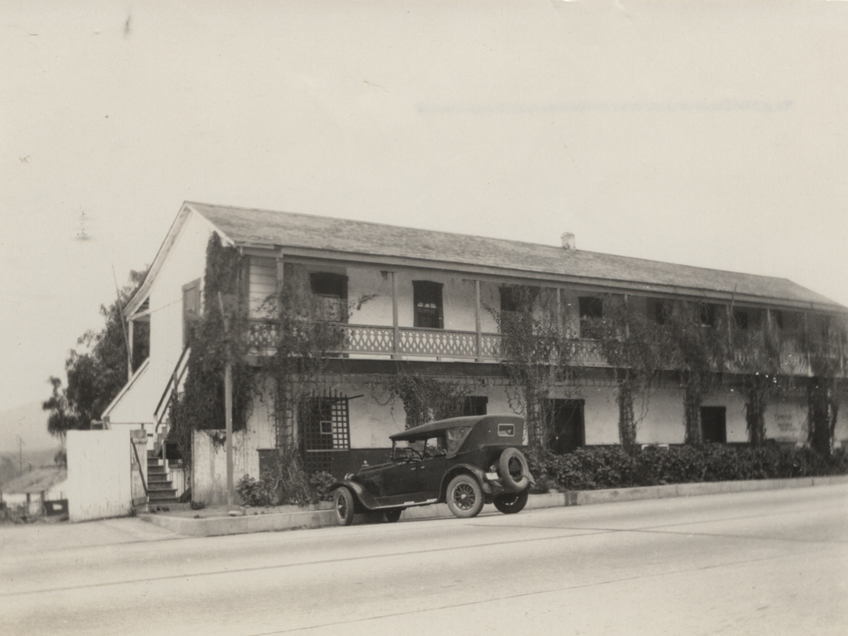 A black vintage car parked in front of an old two-story house with ivy growing on the walls and a balcony.