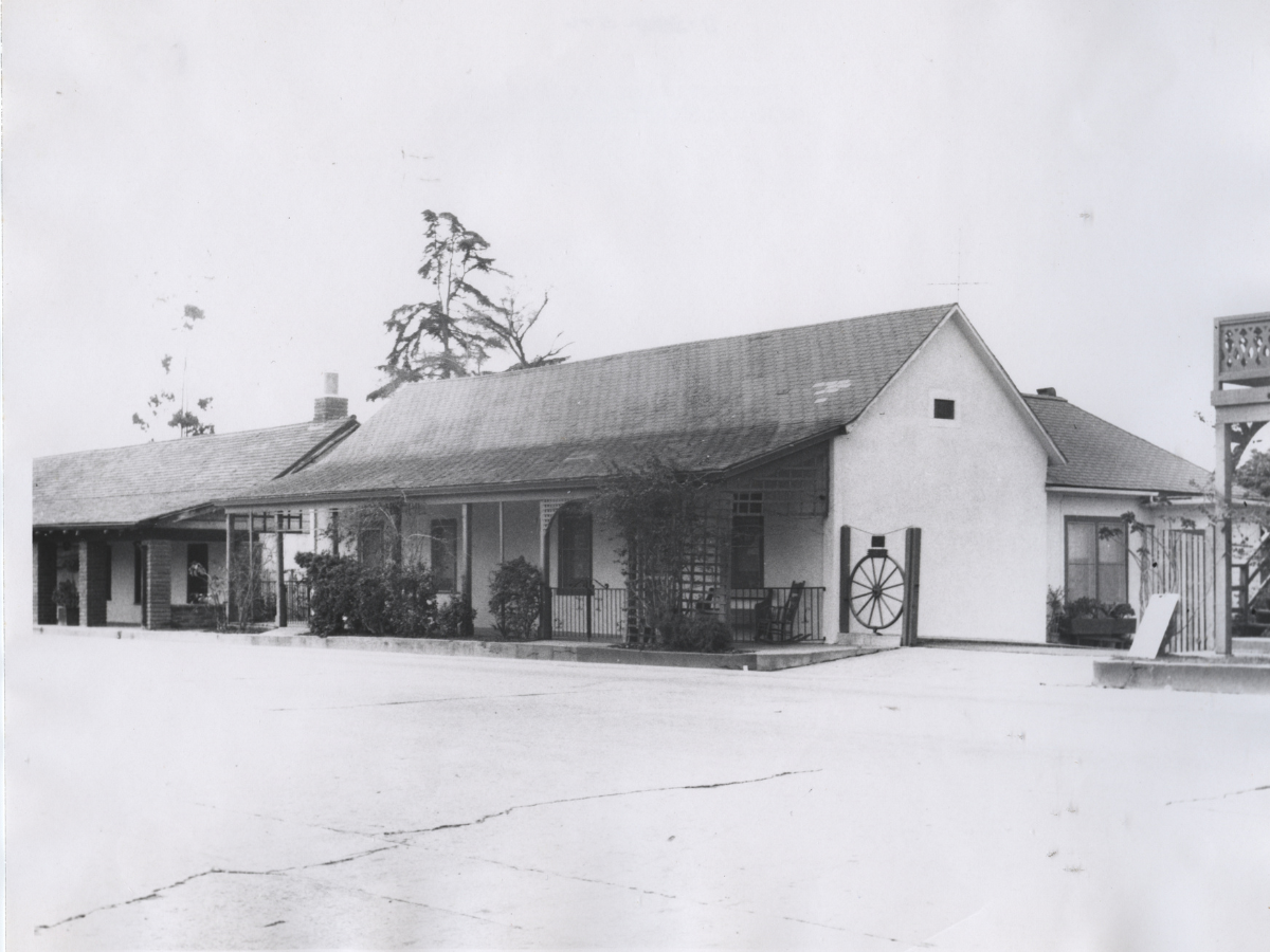 Black and white photo of a house with a sloped roof, small porch, and decorative wagon wheel on the side, with a driveway in the foreground.