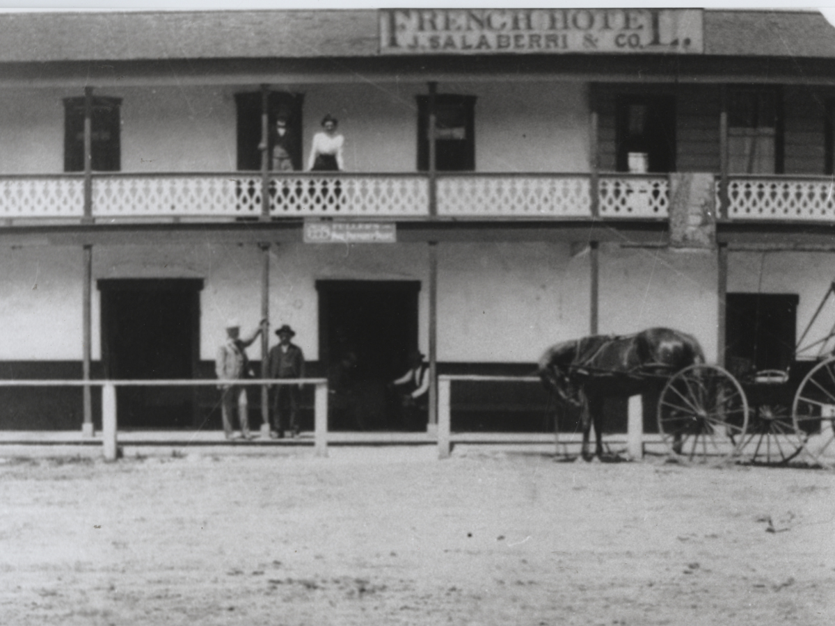 Black and white photo of a two-story building with a balcony, showing several people standing on the balcony and outside the building, and a horse-drawn wagon parked in front.