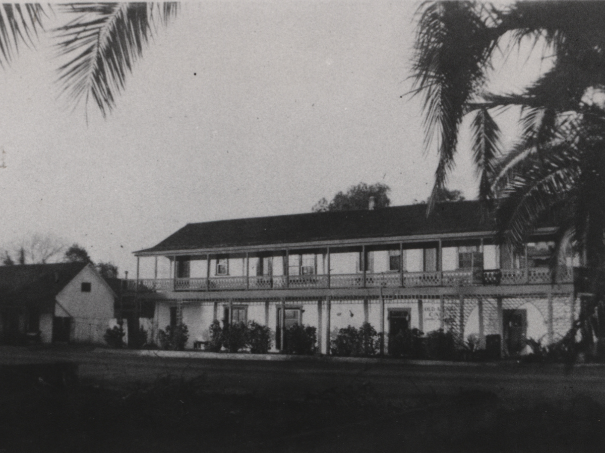 Black and white photo of a two-story house with a wraparound balcony, trees, and a palm tree in the foreground.