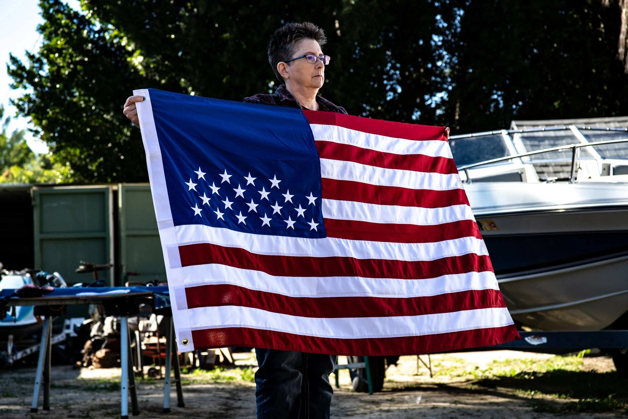 A woman in glasses holds an American flag outdoors, with trees and a boat in the background. There are only 29 stars on the flag signifying the many states where LGBTQ+ Americans lack basic protections.
