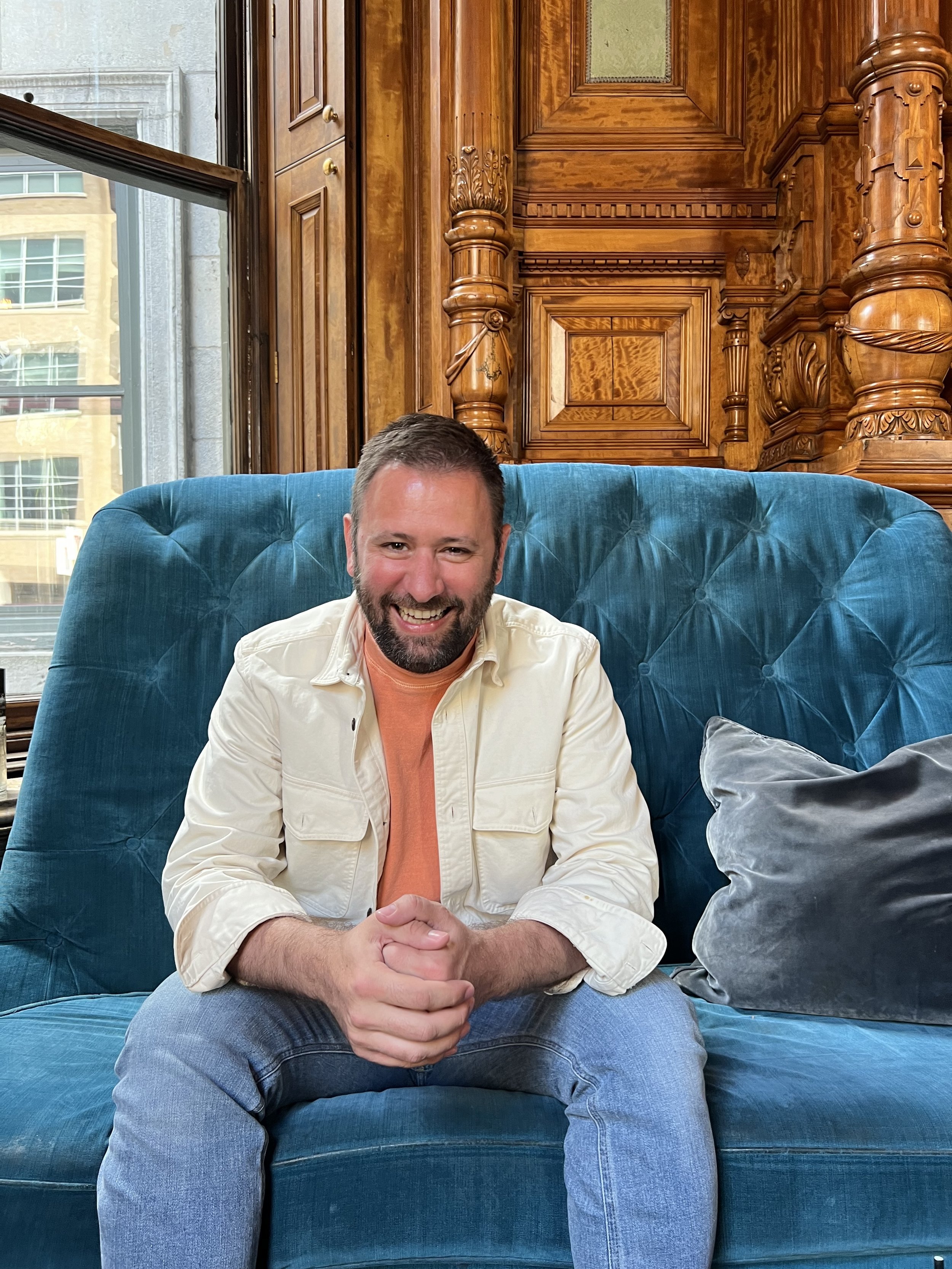 A smiling man named Dom Lowell is looking at the camera. He has short dark hair and a beard, wearing a cream-colored jacket over an orange shirt, sitting on a blue tufted velvet couch in a wood-paneled room with large windows.