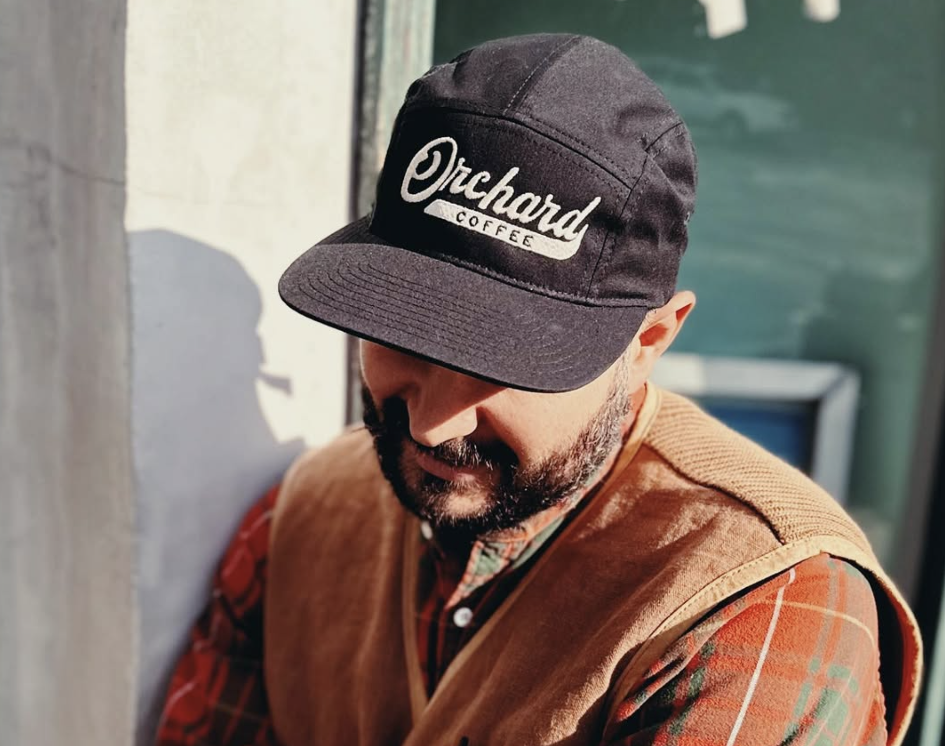 Bearded man wearing a black Orchard Coffee cap, brown vest, and orange plaid shirt outdoors