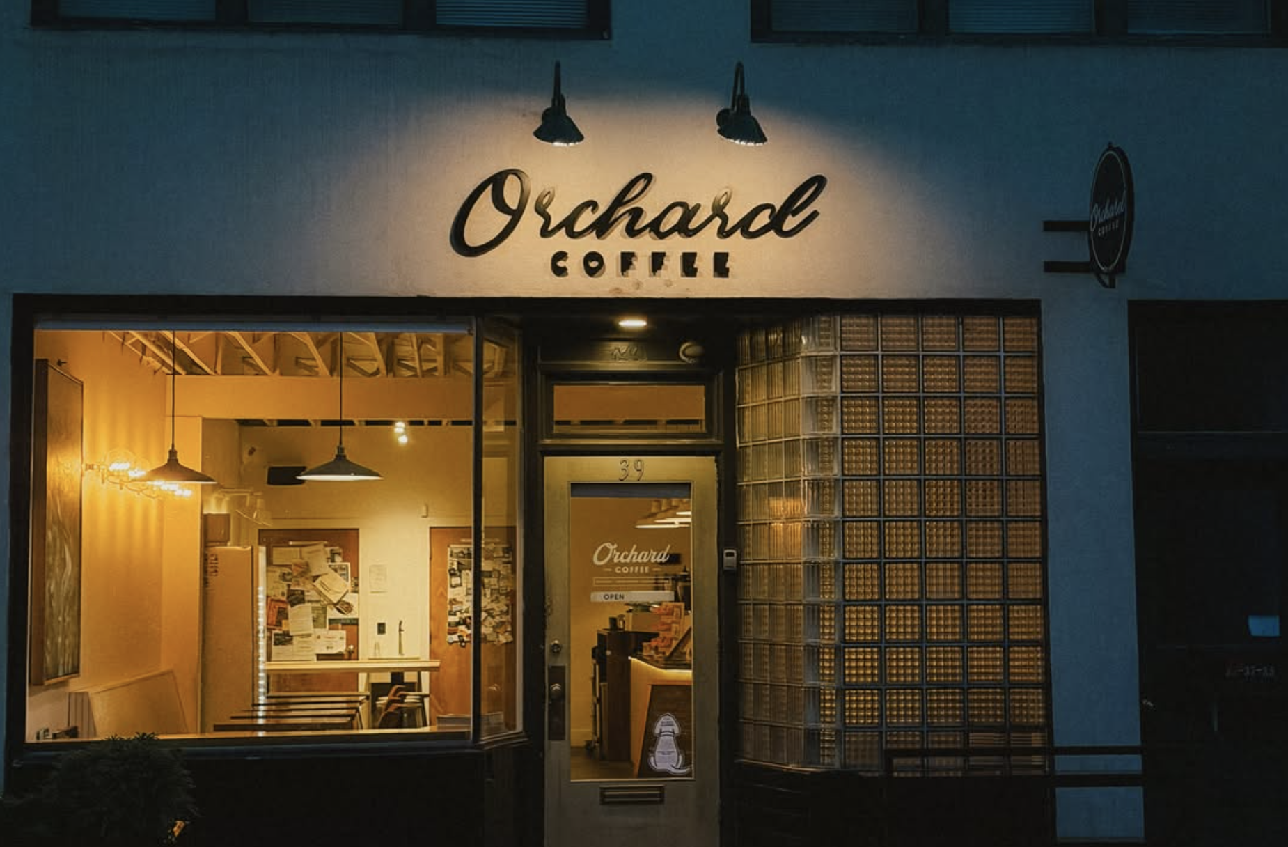 Nighttime exterior of Orchard Coffee shop with illuminated storefront, glass-block wall, and warm interior lighting.