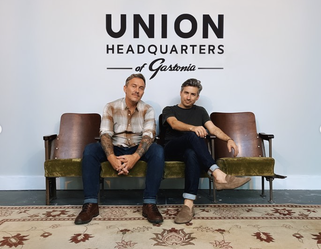 Two men seated in vintage chairs beneath a sign reading “Union Headquarters of Gastonia.”