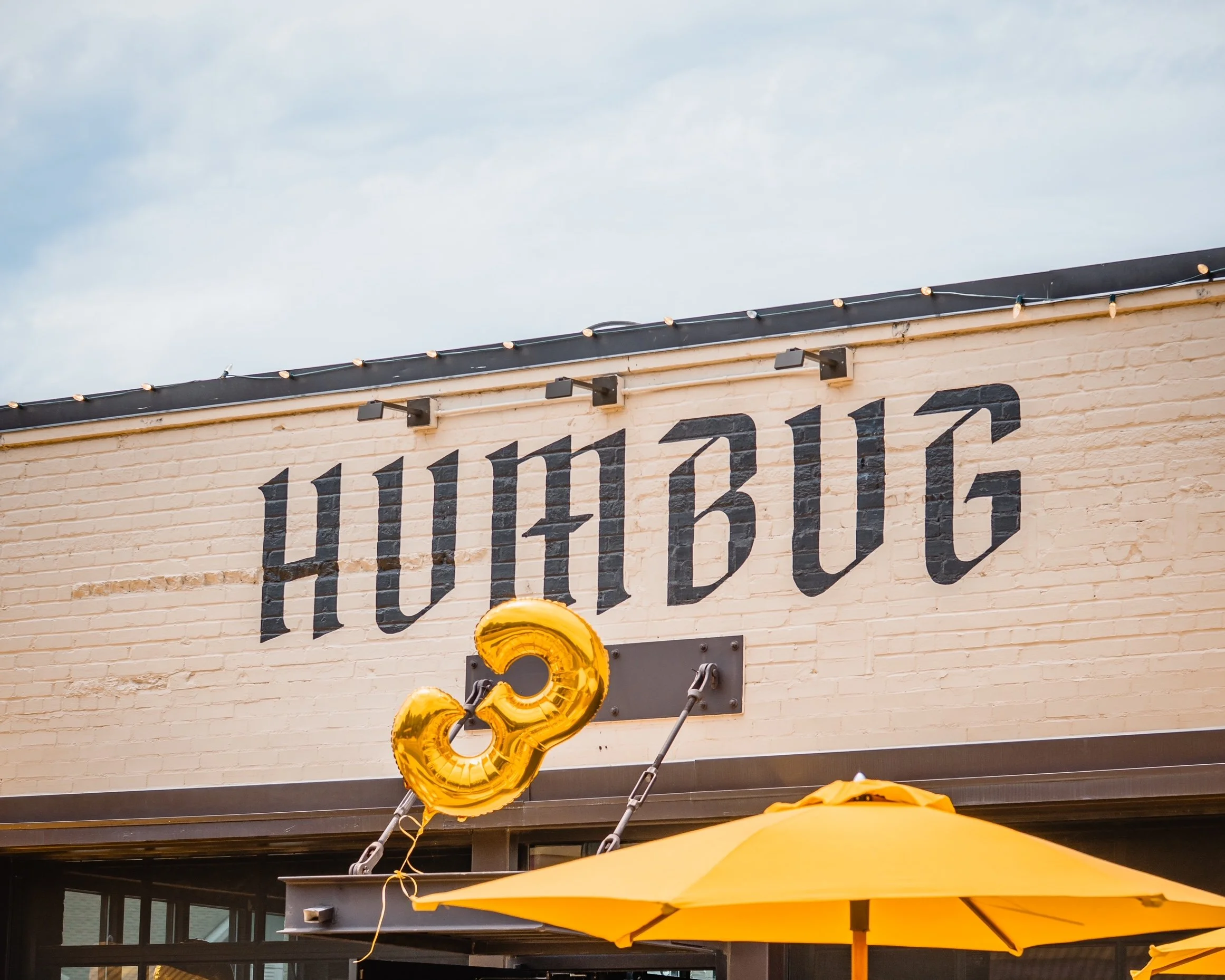Brick building facade with large black “HUMBLE” lettering, string lights, gold number balloons, and yellow patio umbrellas