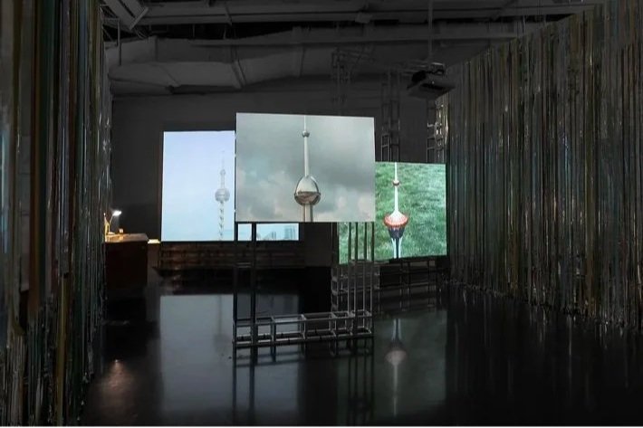 Installation view of a three-channel video installation by Dino Zhang in Danwei Unfolding (2025) at TANK Shanghai, featuring three projections of architectural structures and suspended objects, framed by Nishiko's multicoloured ribbon installation.