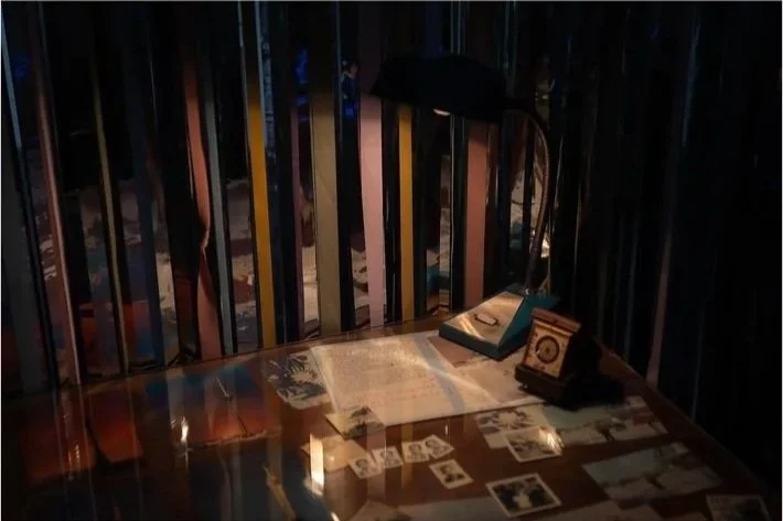 A wooden table with a small desk lamp, a Installation view of archival materials by Dino Zhang in Danwei Unfolding (2025) at TANK Shanghai, featuring archival photographs, a clock, documents and books arranged across a table. Items are illuminated by