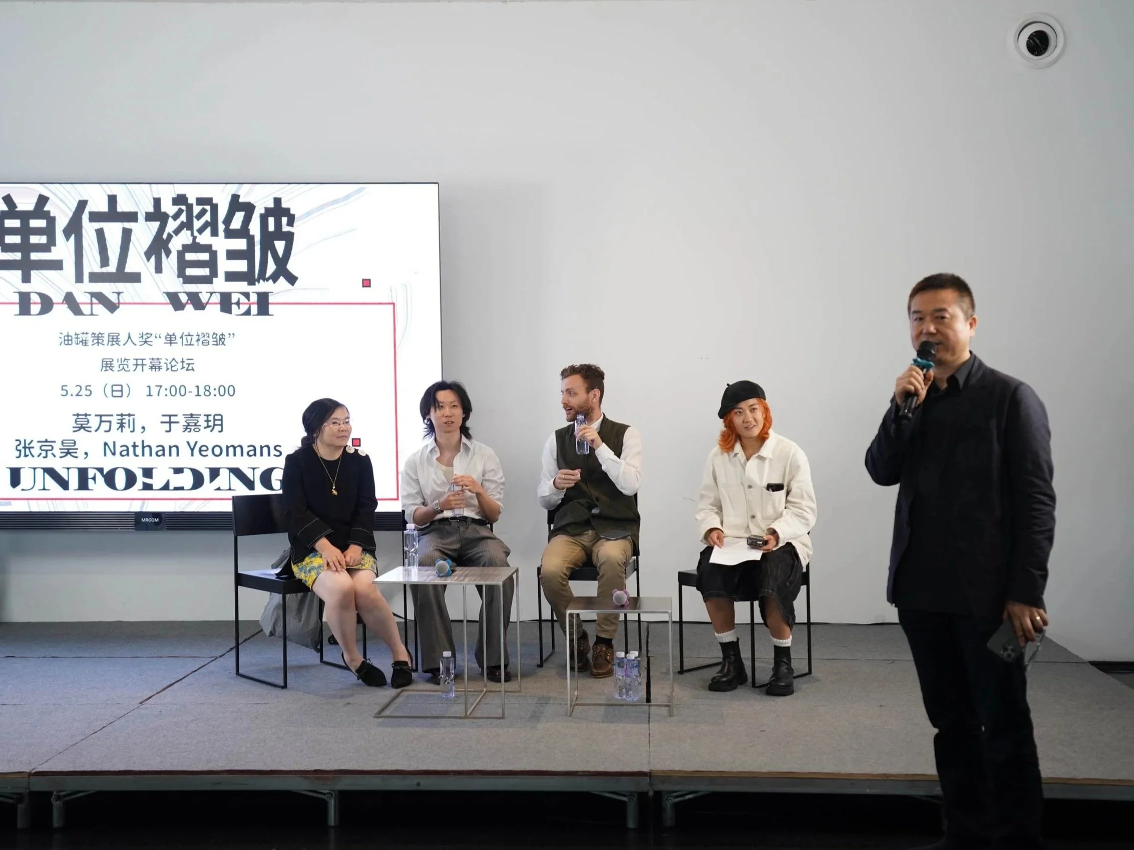 Qiao Zhibing, founder of TANK Shanghai, addressing the audience during a panel discussion accompanying Danwei Unfolding (2025), with invited speakers seated on stage and the exhibition's event programme displayed behind them.