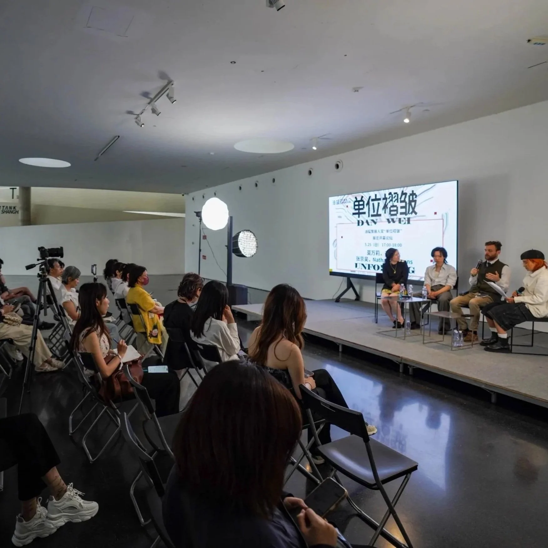 A panel discussion and symposium for TANK Shanghai's Danwei Unfolding, taking place at a conference or seminar with people sitting on a stage and an audience watching. There is a large screen displaying event details in the background. Gues speakers 