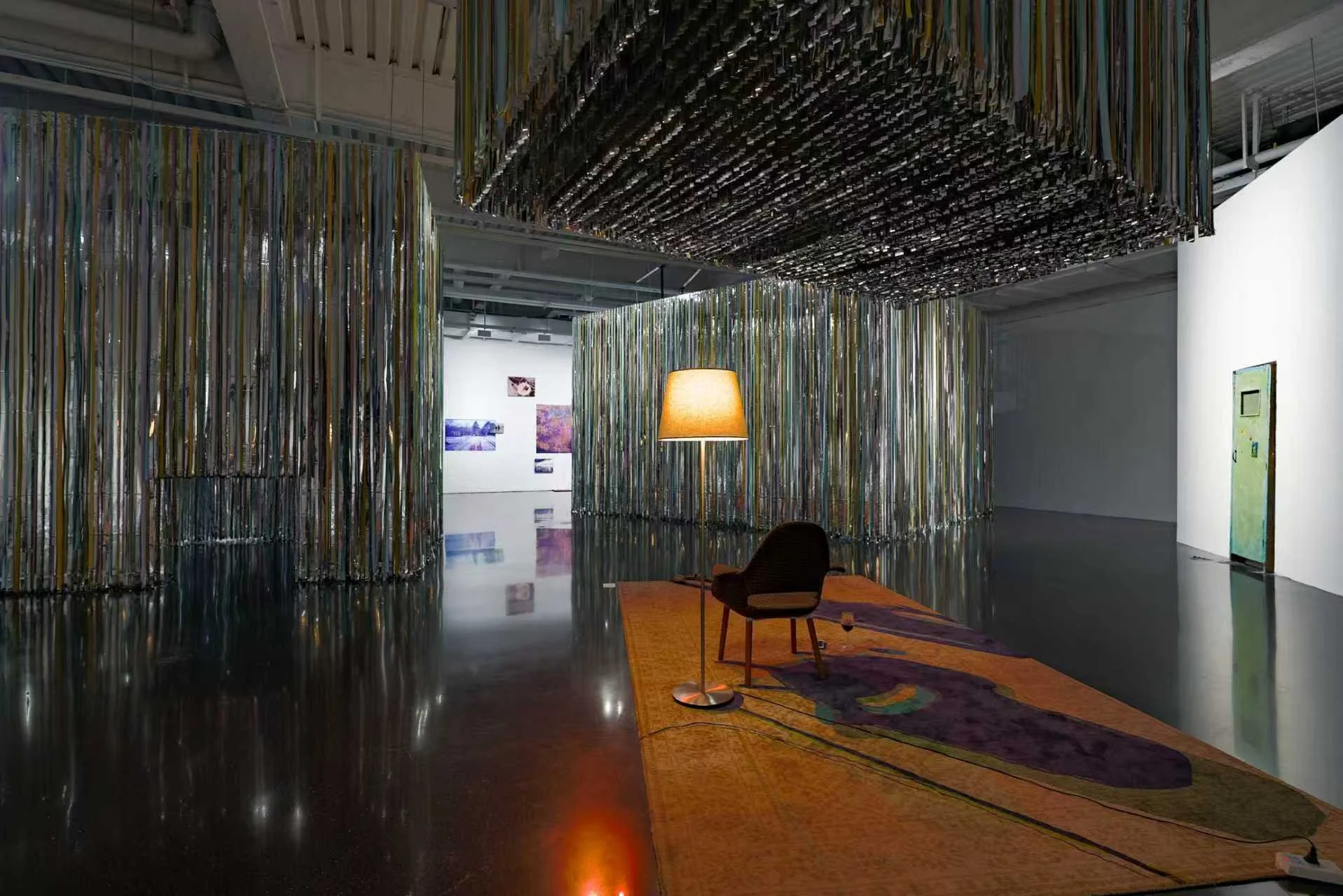 Installation view of Danwei Unfolding (2025) at TANK Shanghai, featuring Xing Danwen's installation with a chair, floor lamp and patterned carpet, alongside suspended metallic ribbons. Works by Zheng Tianyi are visible in the background, with a paint