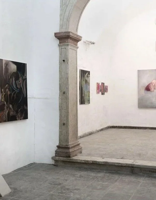 Art gallery with paintings on white walls and an arched stone pillar in the center of the room.