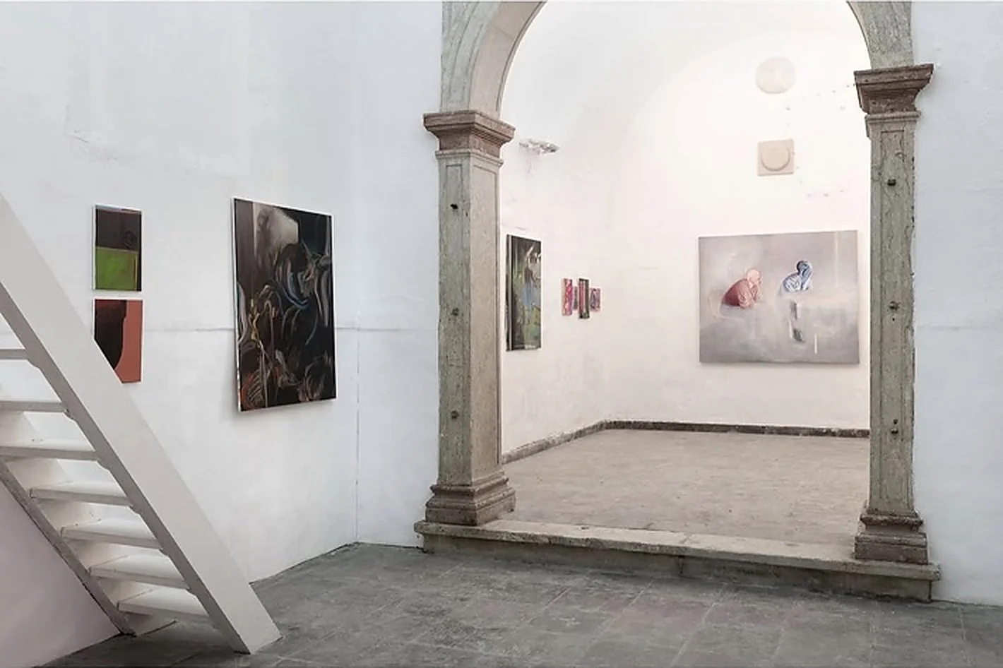 Installation view of Déjà Vu (2024) at Dialogue Gallery, Lisbon, showing a dark painting by Tom Plehn in the foreground and works by Luca Japkinas and other artists beyond the stone column.